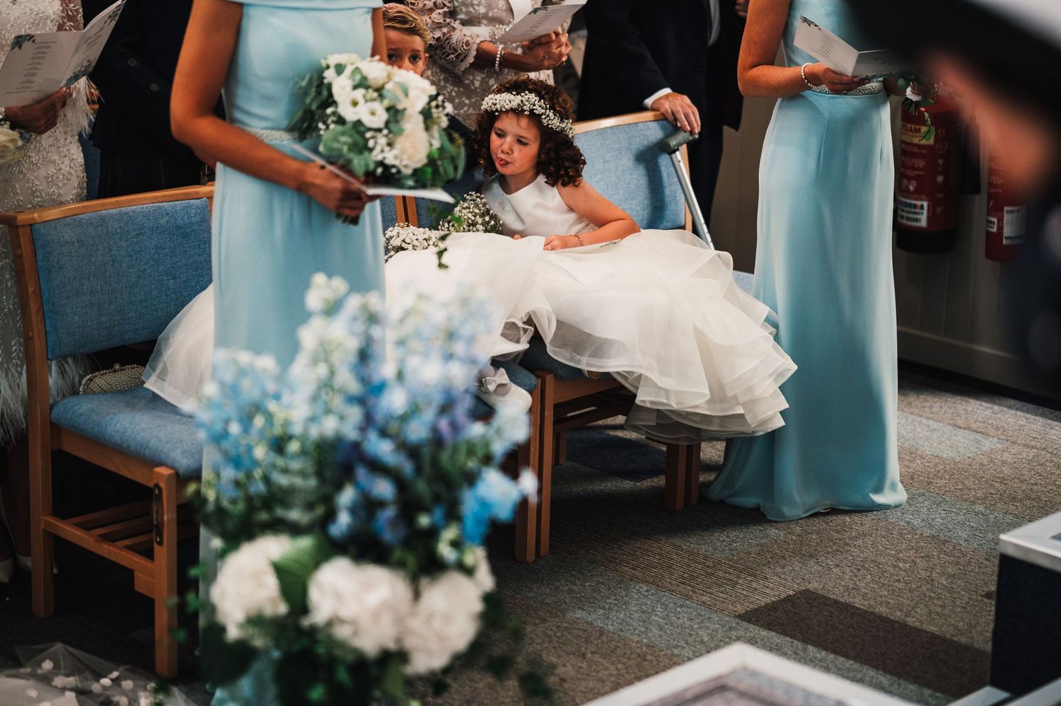 A flower girl in white dress sits between bridesmaids wearing light blue dresses with floral bouquets.