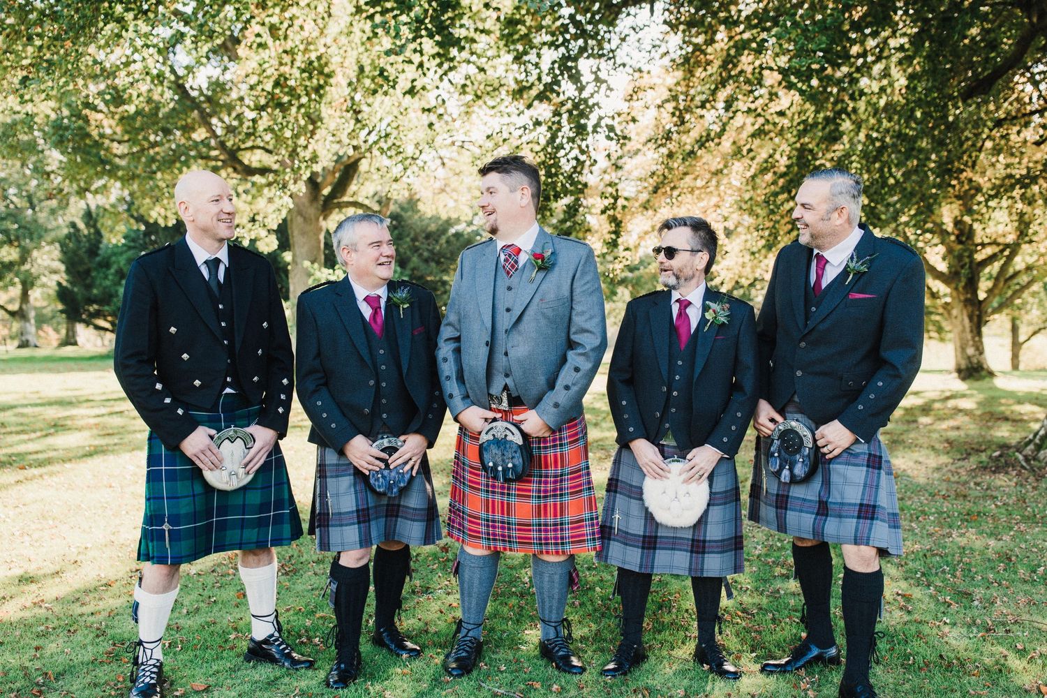 Group of five men wearing traditional Scottish kilts standing together outdoors.