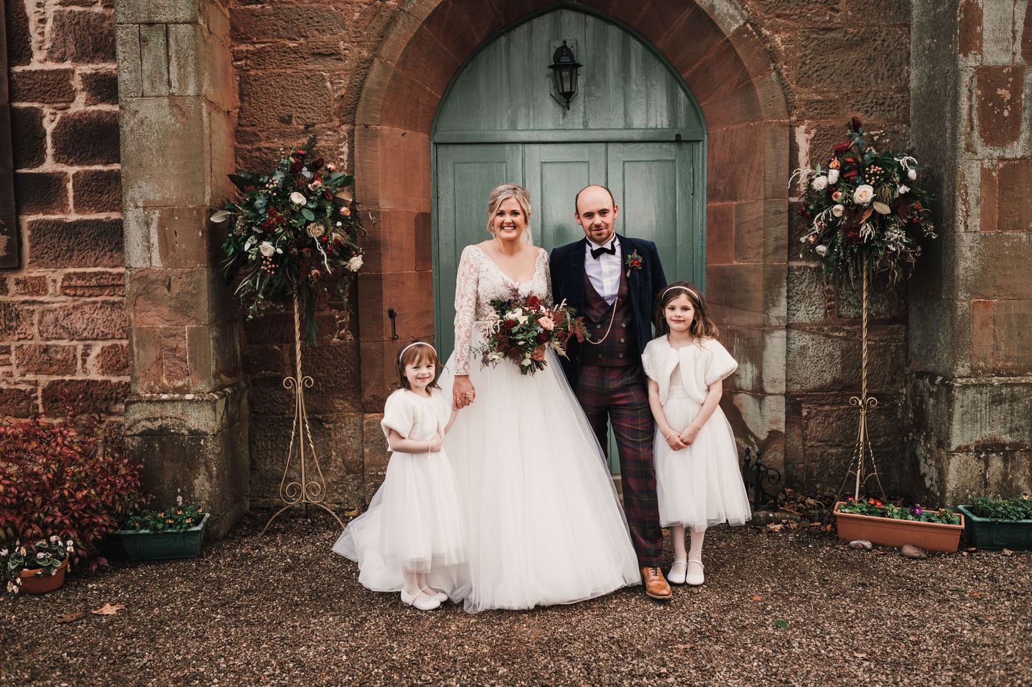 Wedding couple with flower girls pose in front of decorated church entrance.