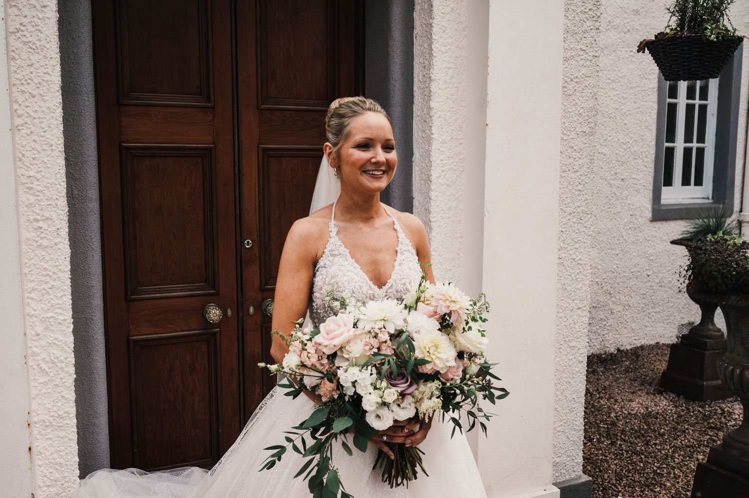 Bride in white V-neck gown holding lush bouquet of roses and greenery by wooden door.