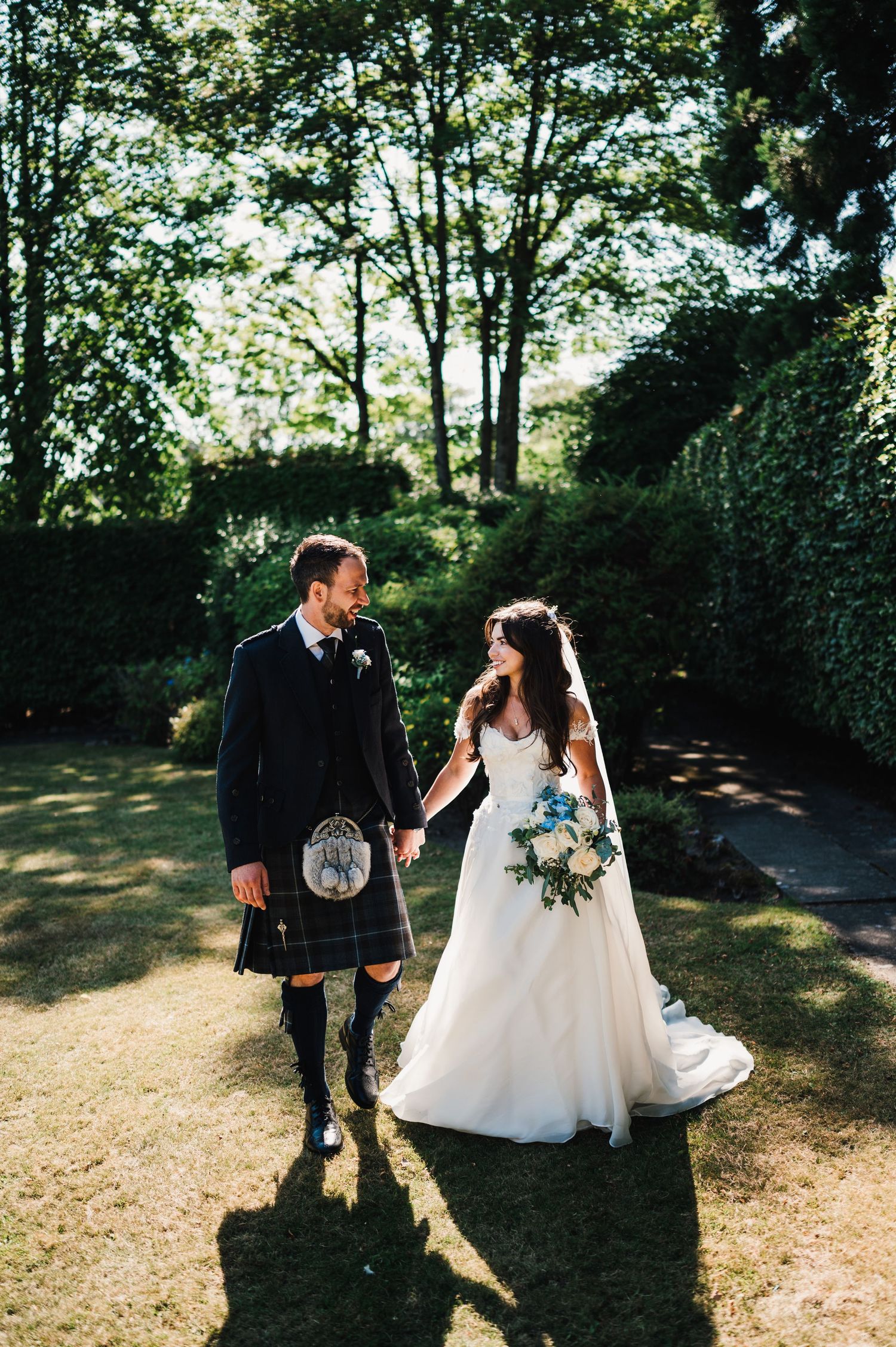 Wedding couple walks hand in hand through sunny garden, groom wearing traditional Scottish kilt.