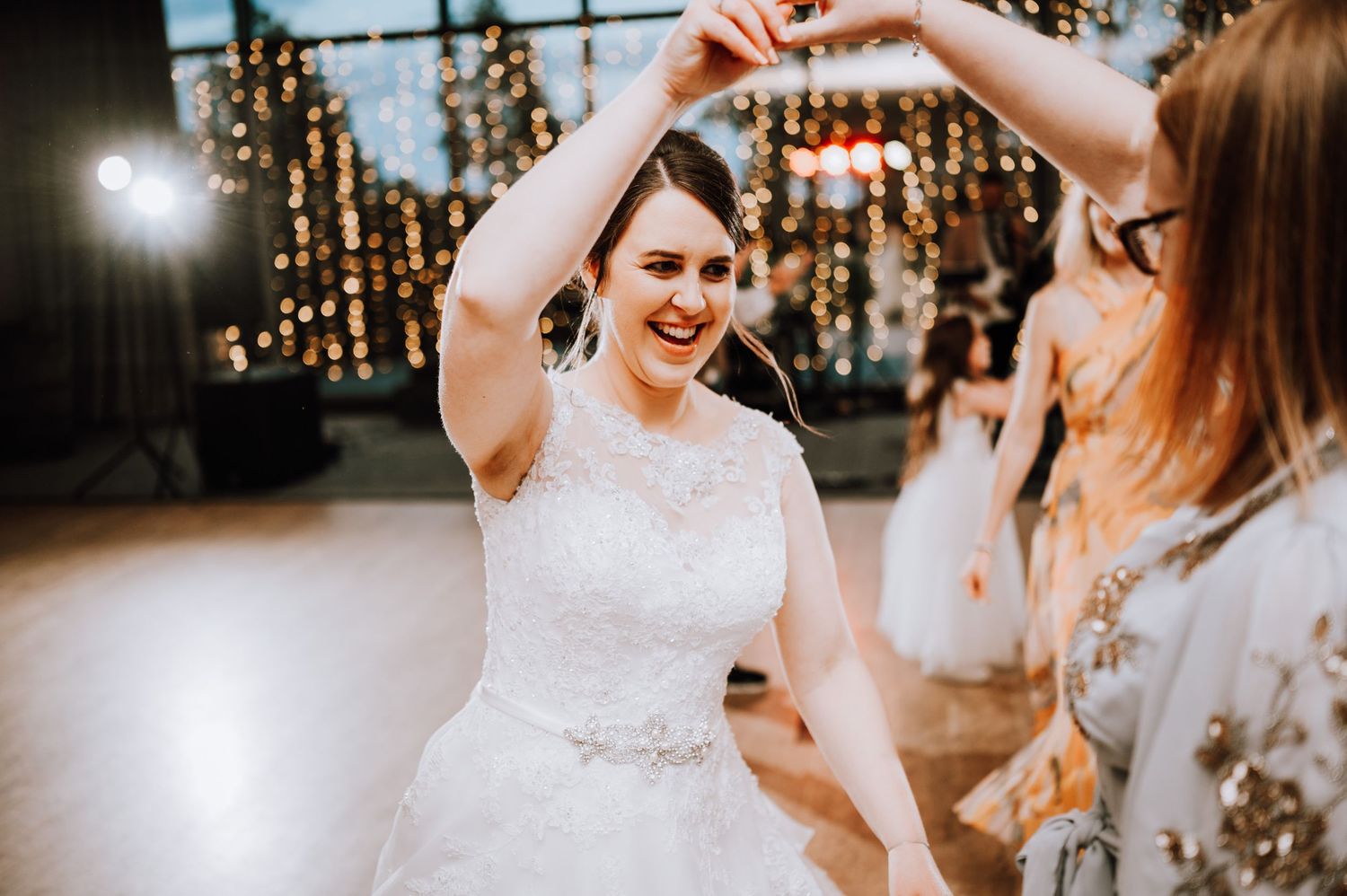 A joyful person in a white wedding dress dances under twinkling lights at a wedding reception.