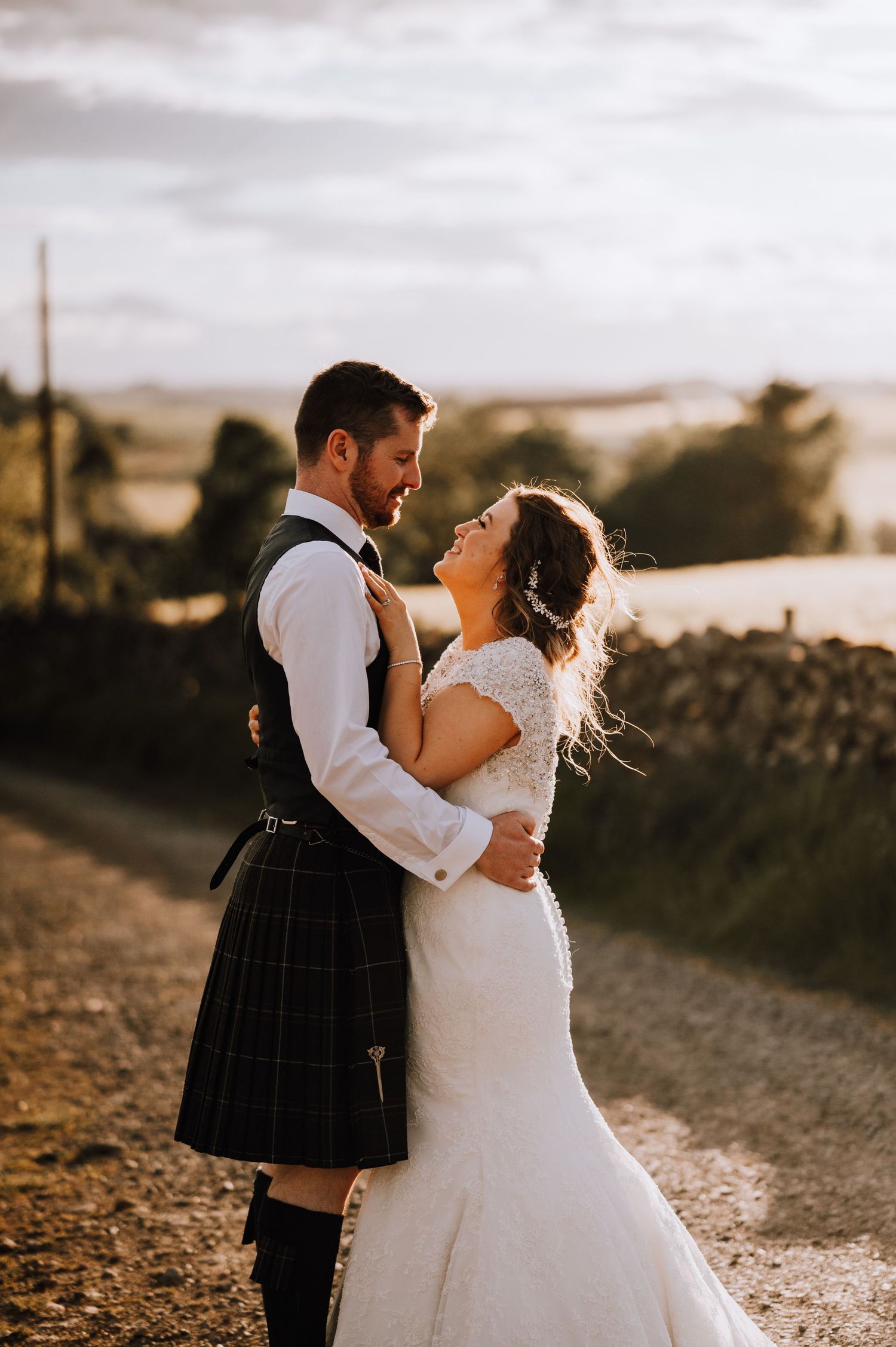 Wedding couple in traditional Scottish attire pose on a countryside path at sunset.