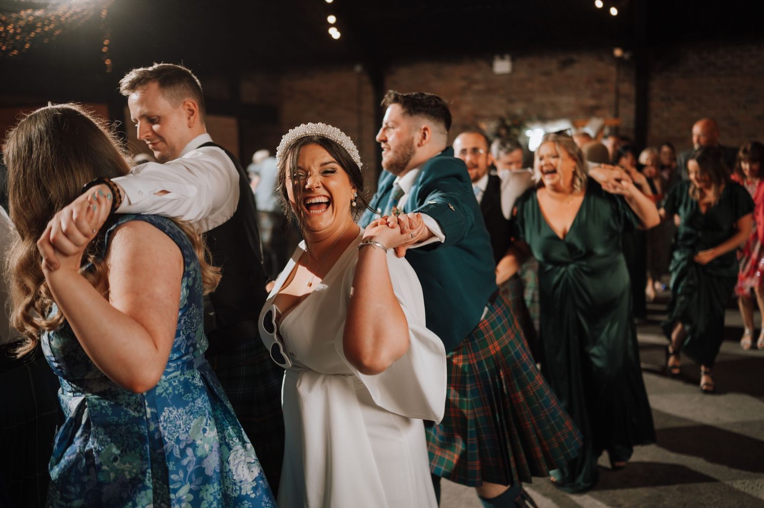 Wedding guests laugh and dance together at a lively indoor reception with string lights.