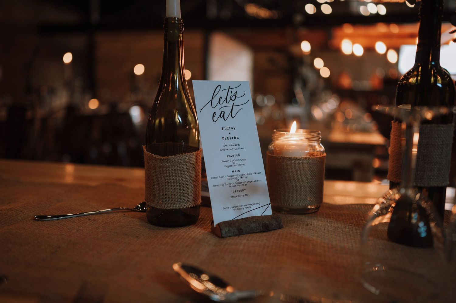 Menu card and wine bottle on rustic wooden table with ambient candlelight in restaurant setting.