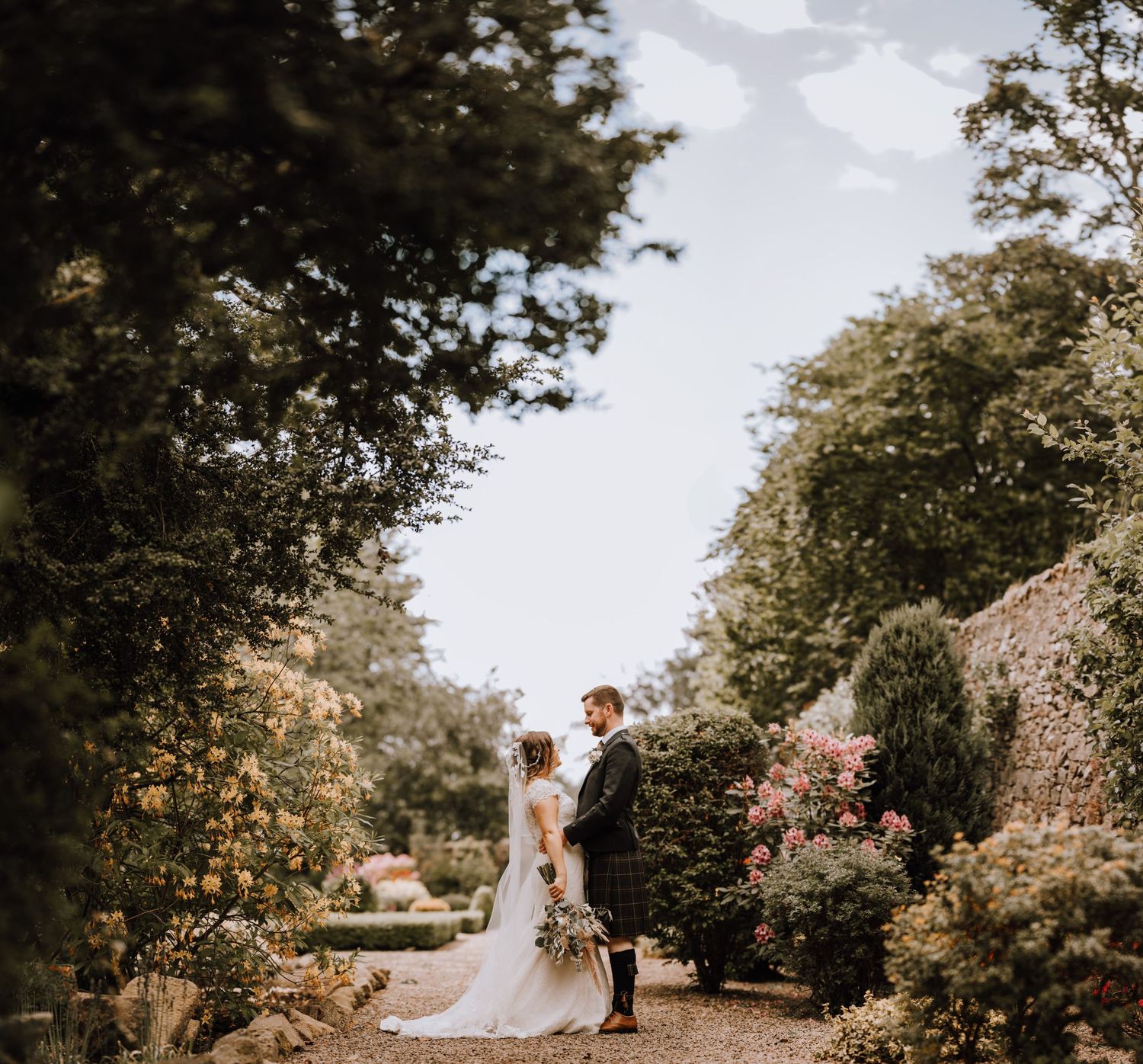 A romantic garden wedding scene with a couple standing on a winding path surrounded by lush greenery.