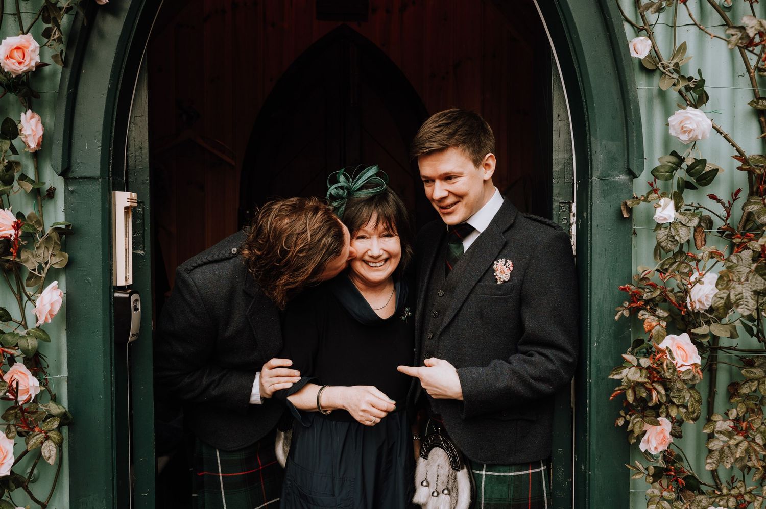 Three people share a joyful moment in a green archway decorated with roses.