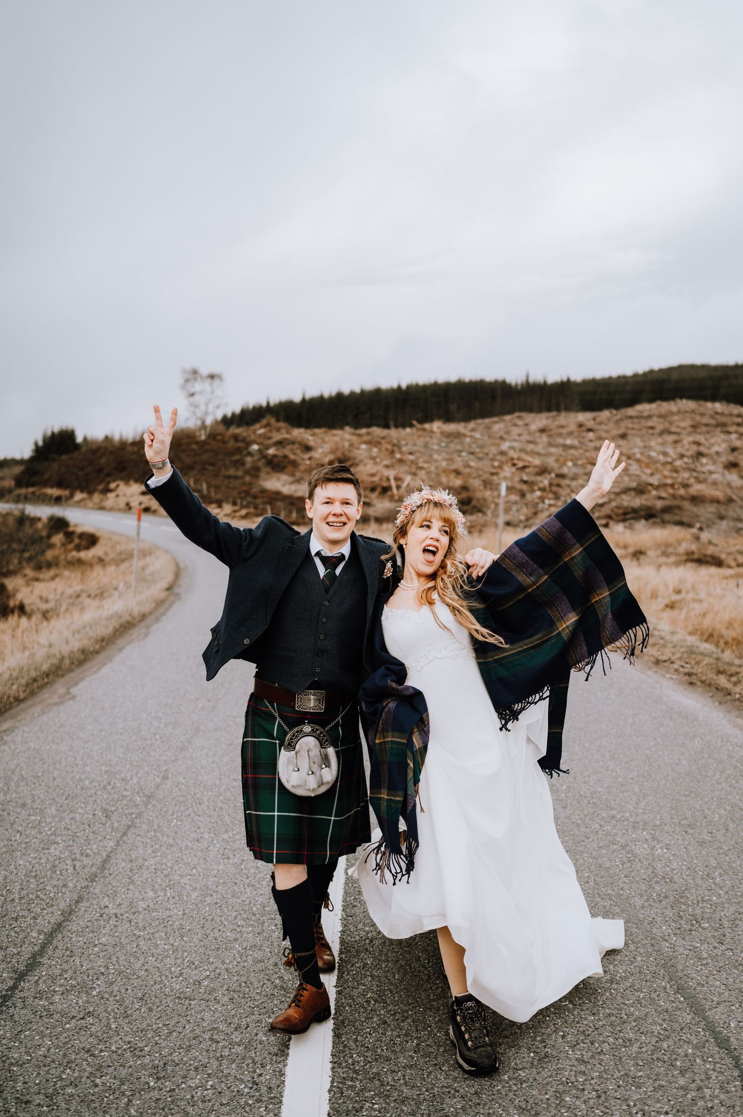Newlyweds celebrate joyfully on mountain road wearing traditional Scottish wedding attire.