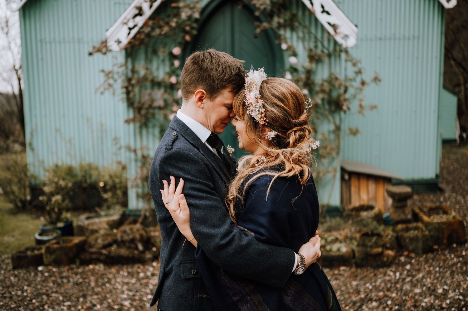 Couple shares intimate moment wrapped in black shawl outside mint green chapel on wedding day.