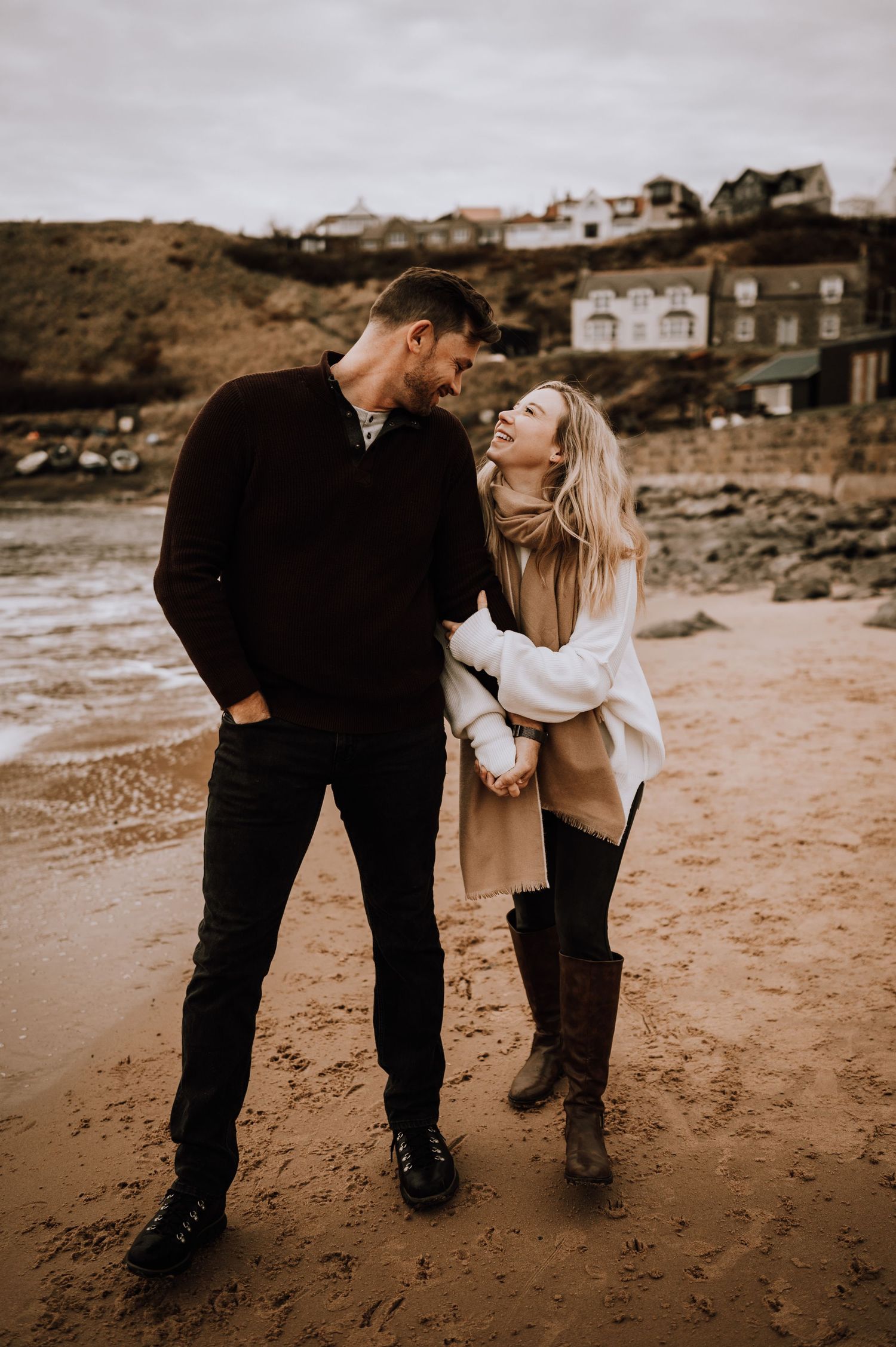 A romantic couple sharing an intimate moment while walking on a sandy beach with houses on the cliff.