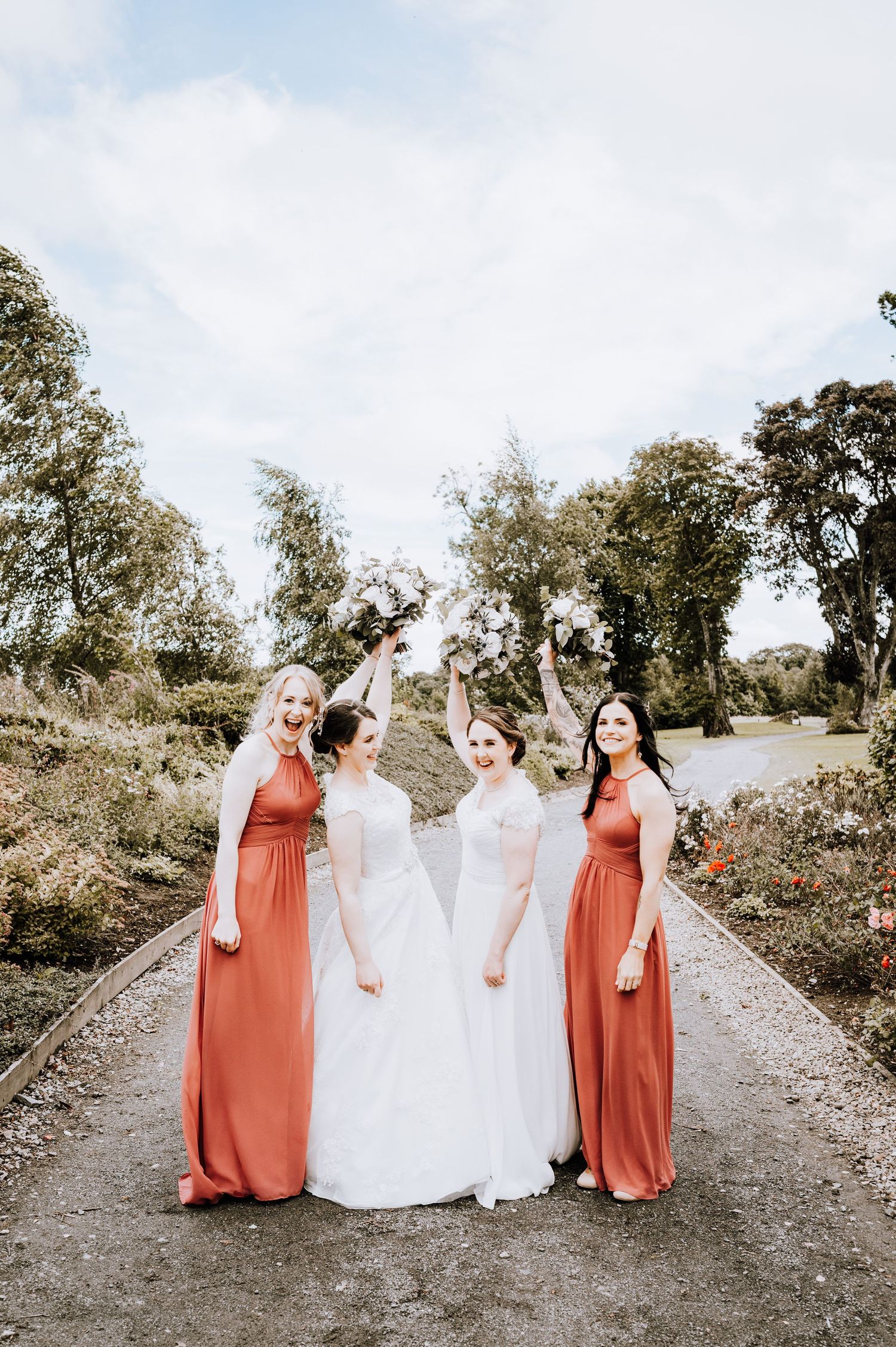 Wedding group portrait on a country path with bridesmaids in coral dresses and white bouquets.