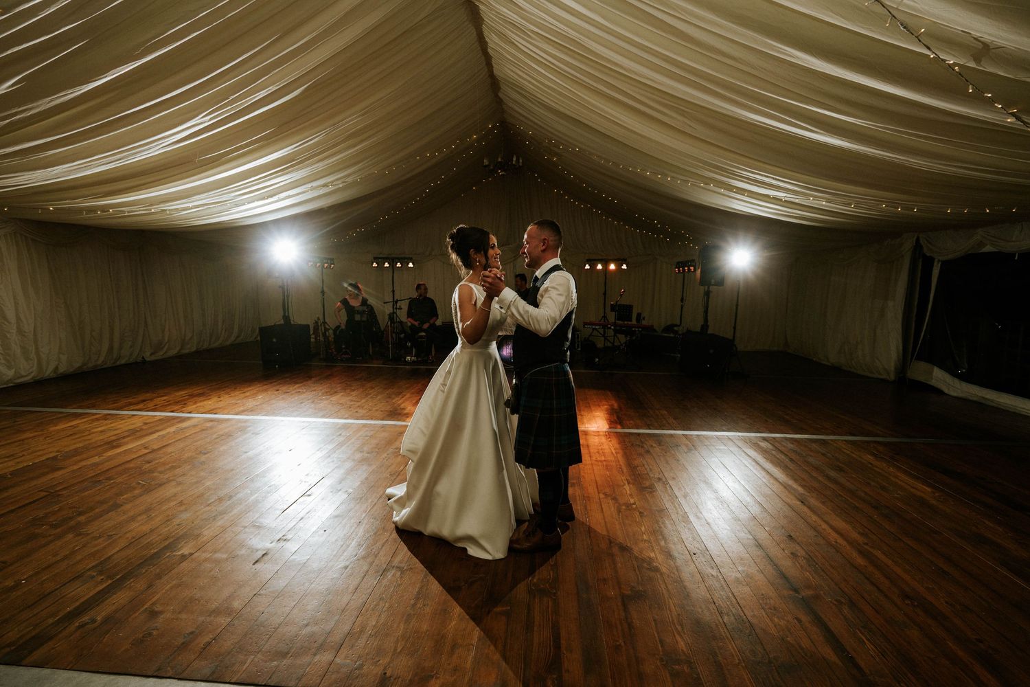 Couples dancing together on a wooden dance floor under draped fabric ceiling with string lights at a wedding reception.