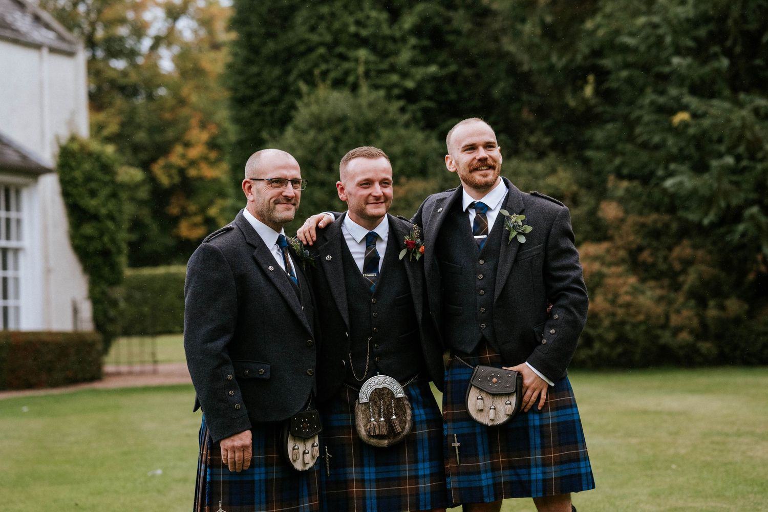 Groomsmen in traditional Scottish kilts and formal wear pose on a lawn.