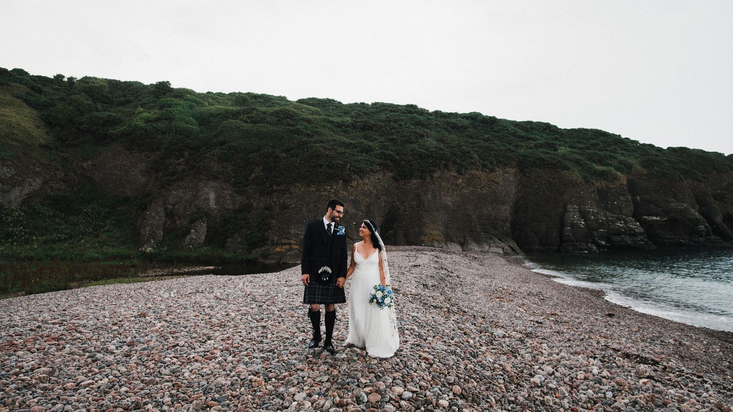A wedding couple walks along a dramatic pebble beach with towering cliffs behind them.