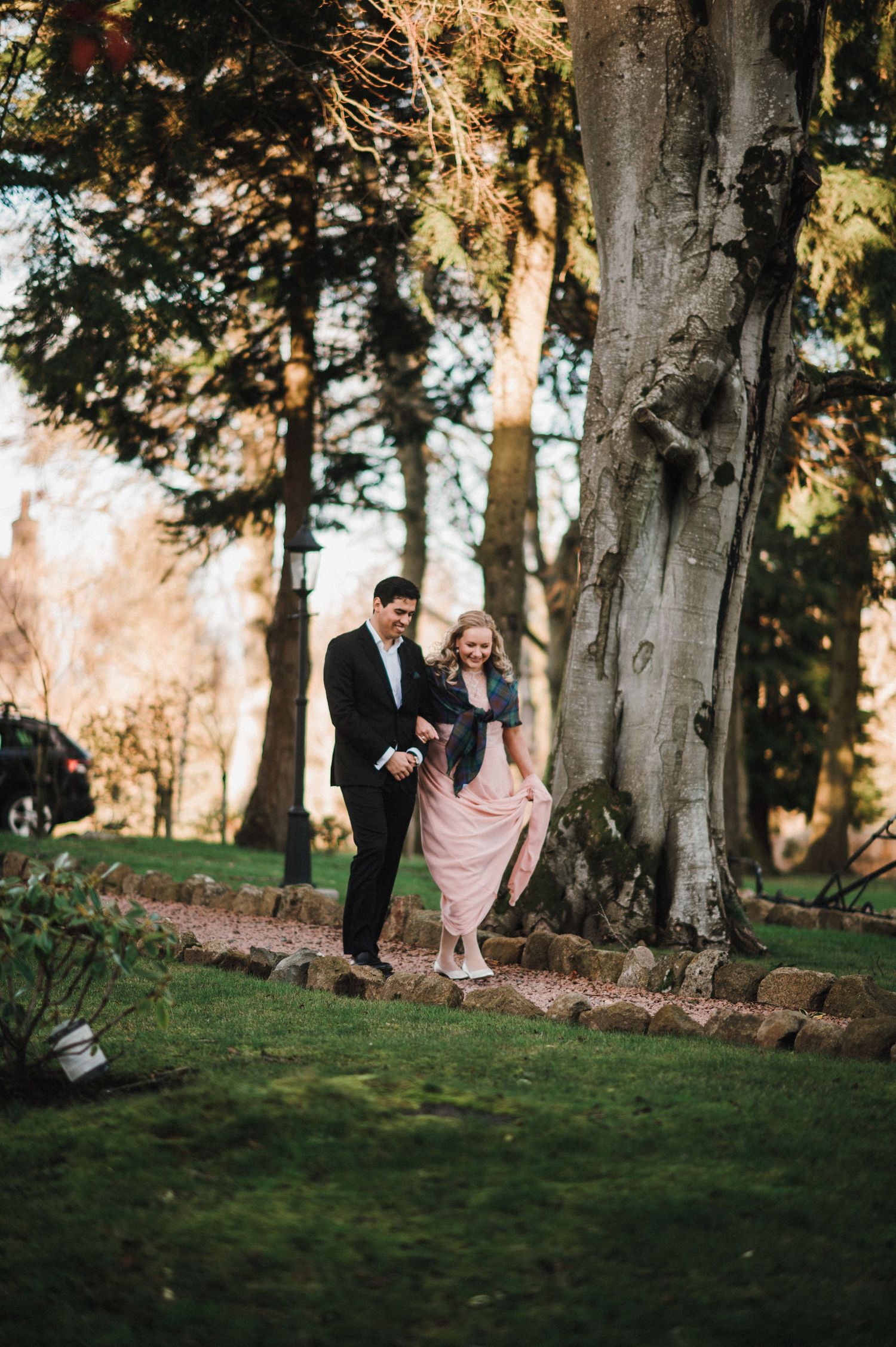A couple walks together on a garden path surrounded by tall trees during golden hour.