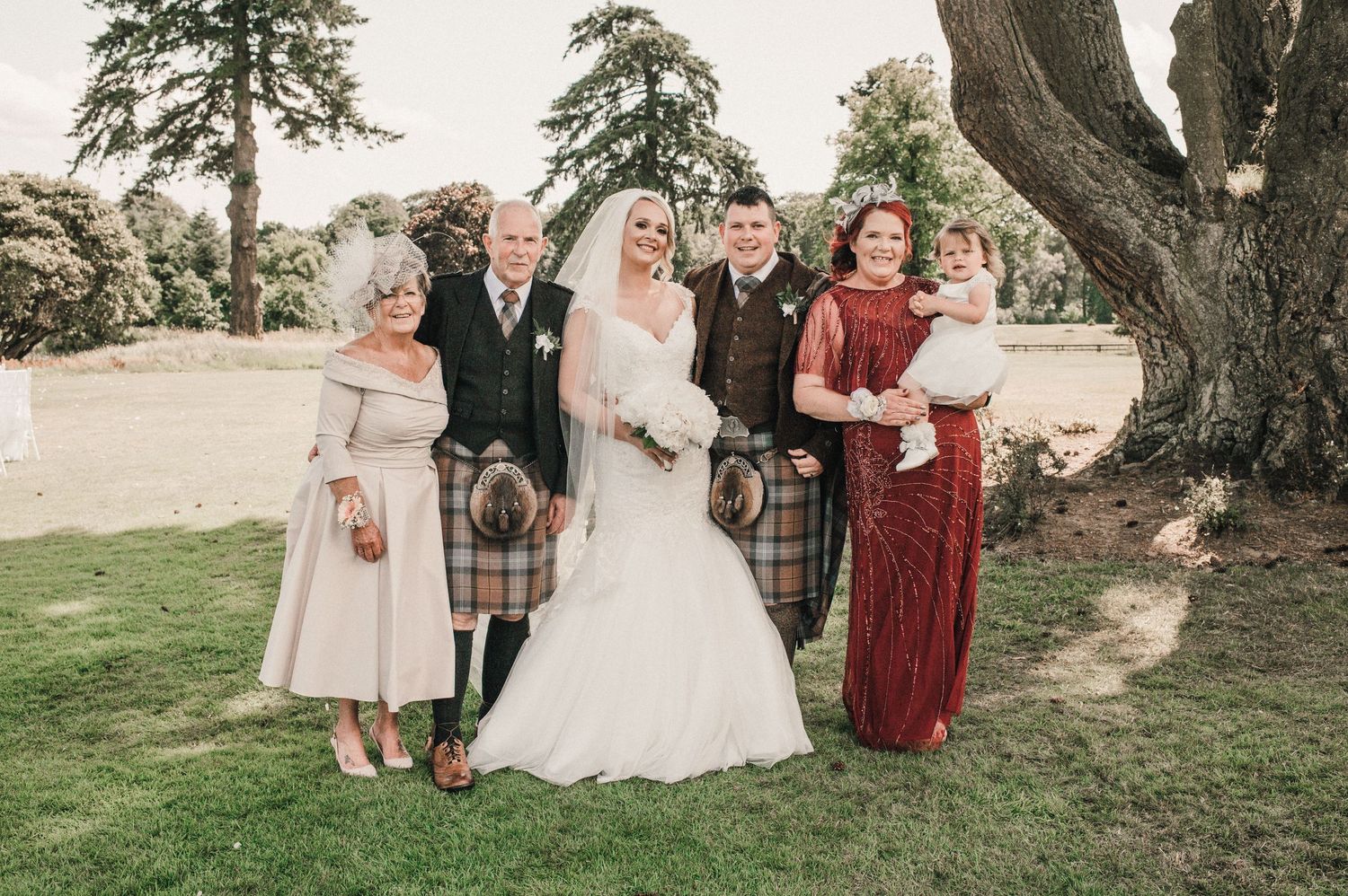 Wedding party in traditional Scottish attire and formal wear pose under ancient tree.