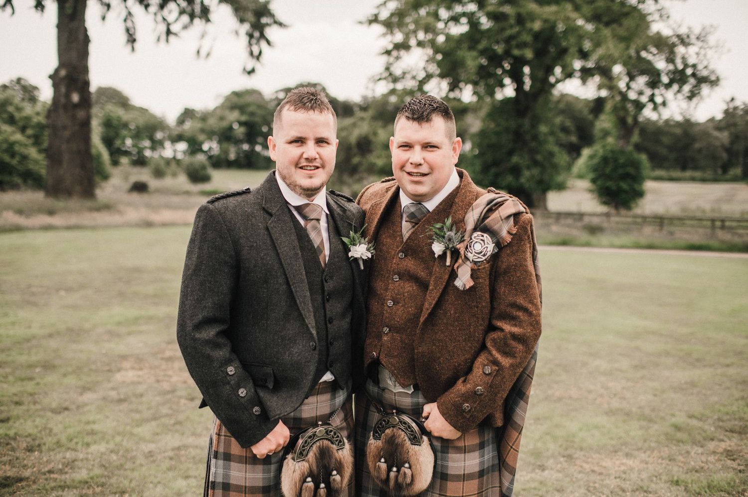 Two people in kilts and formal jackets pose together in a grassy field.