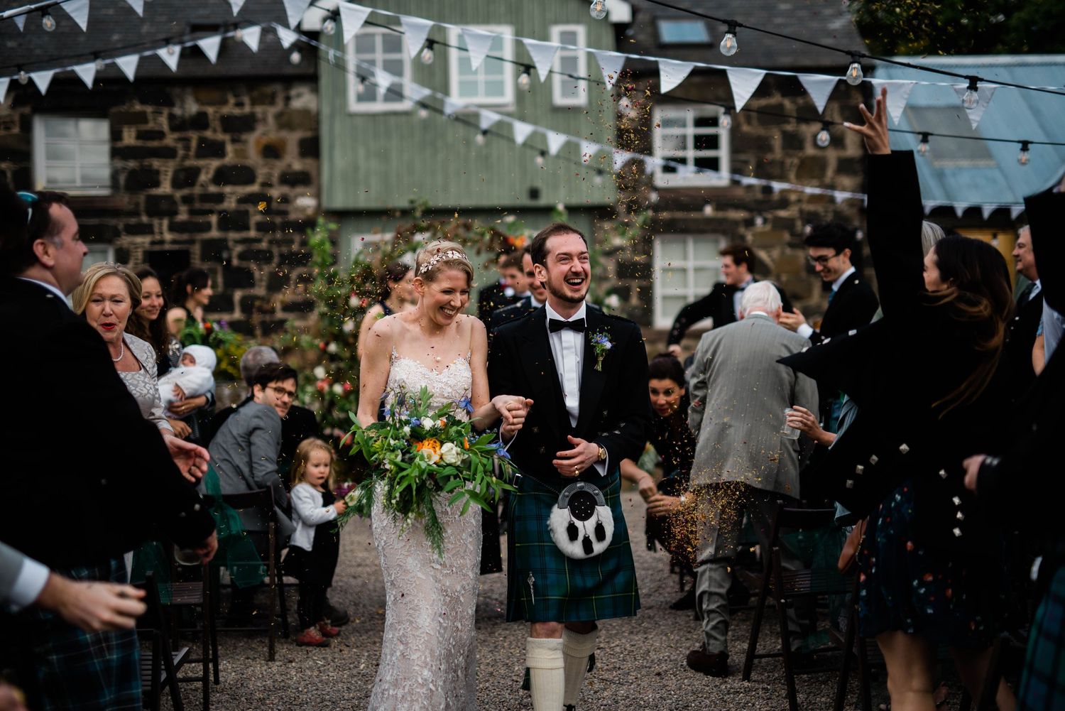 A festive outdoor wedding celebration with white bunting decorations and traditional Scottish kilts.