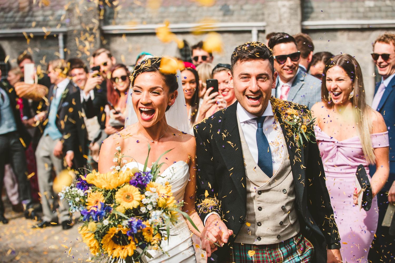 Joyful couple celebrating with confetti at outdoor wedding while wearing traditional Scottish attire and holding sunflowers.