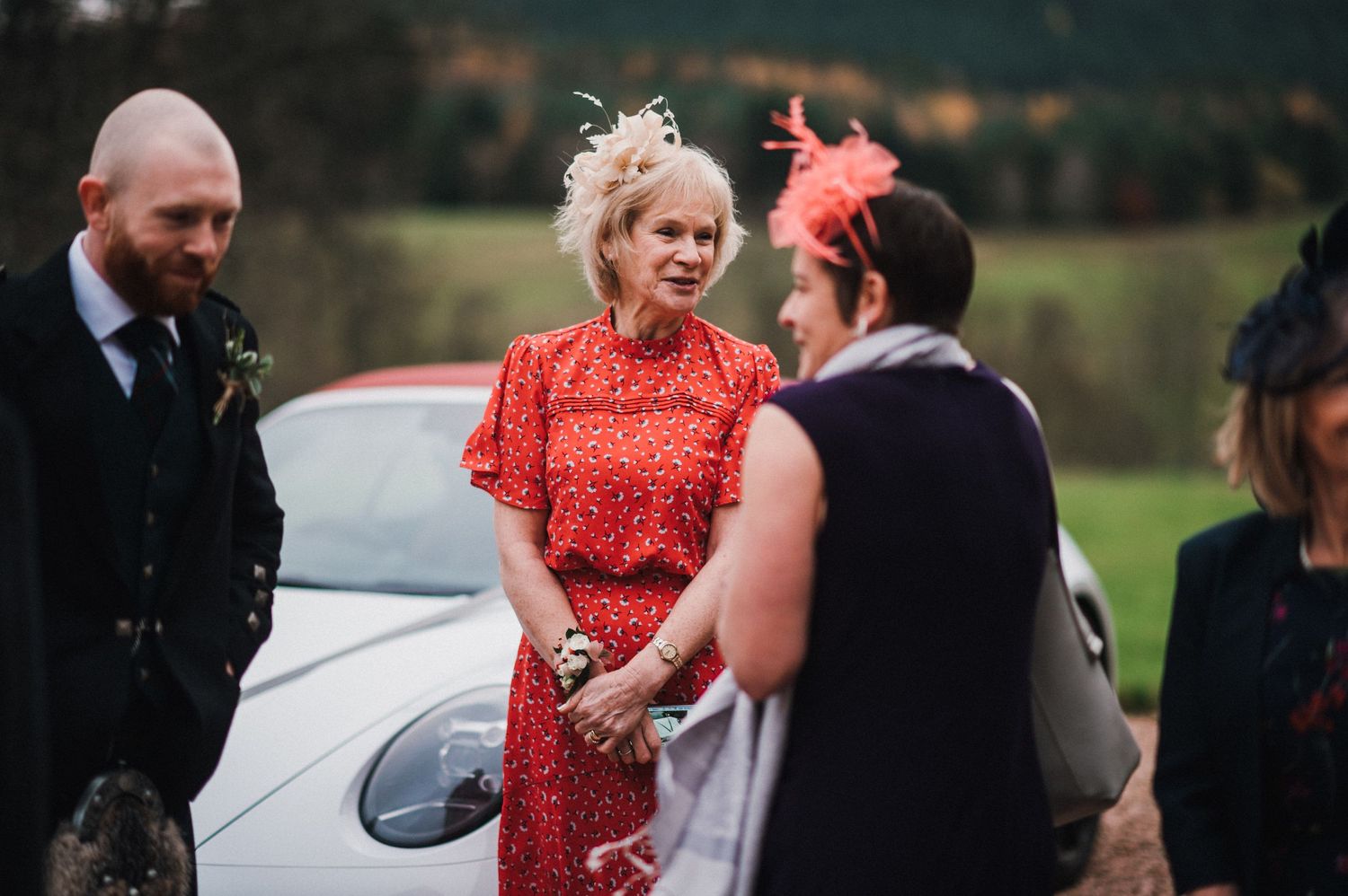 Wedding guests in formal attire and colorful fascinators chat near parked car.