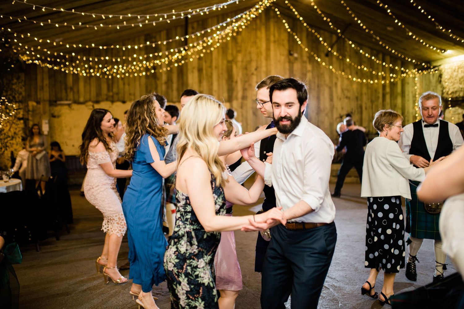 Wedding guests dance and celebrate beneath warm string lights at an indoor reception.