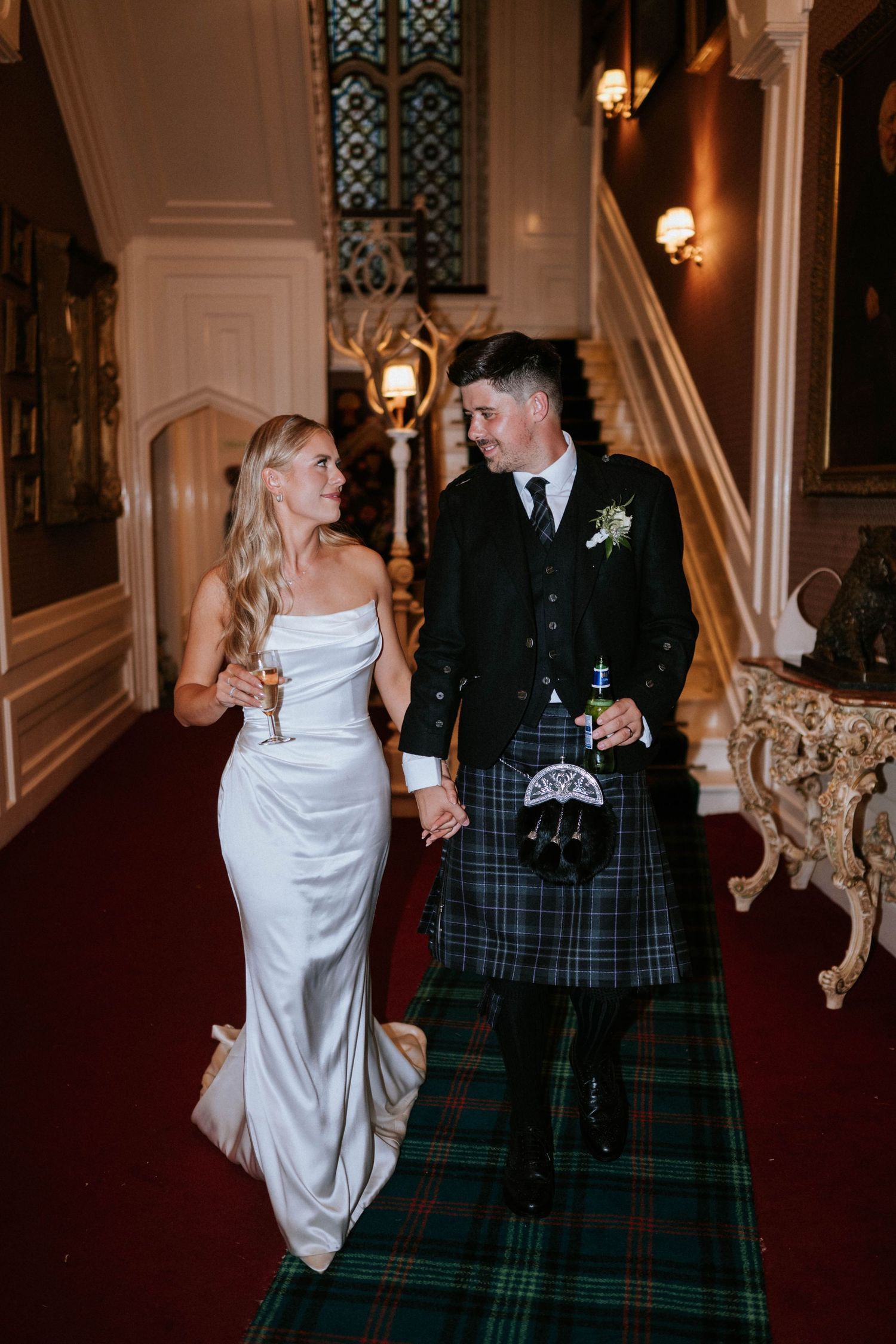 Couple in wedding attire walks down elegant hallway with traditional Scottish elements.
