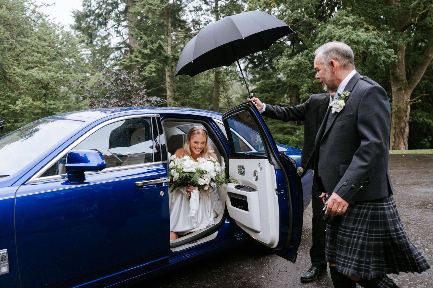 Wedding party arrives in sleek blue vintage car on rainy day with umbrellas.