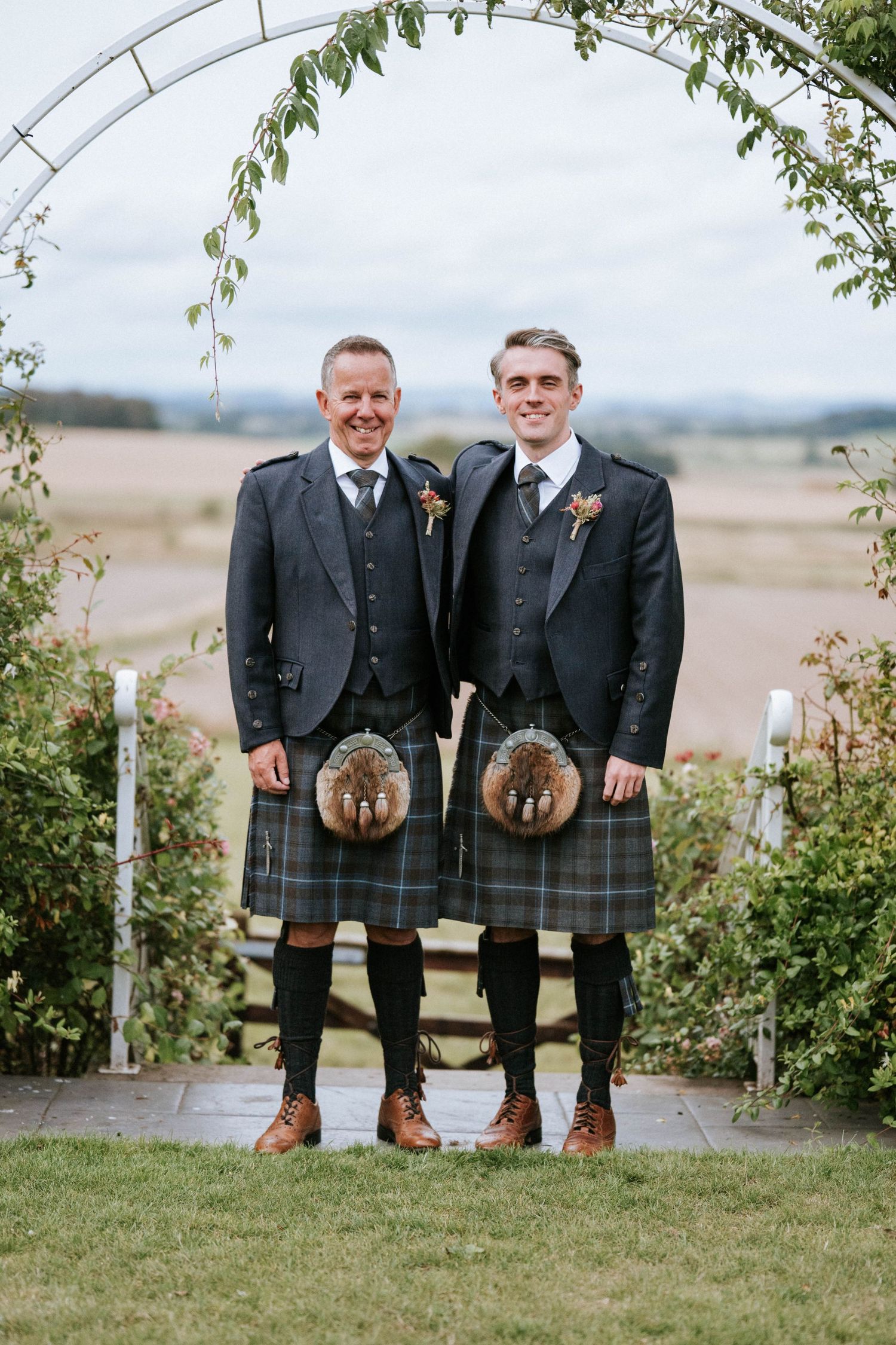 Two men in traditional Scottish kilts and formal attire standing under floral arch.