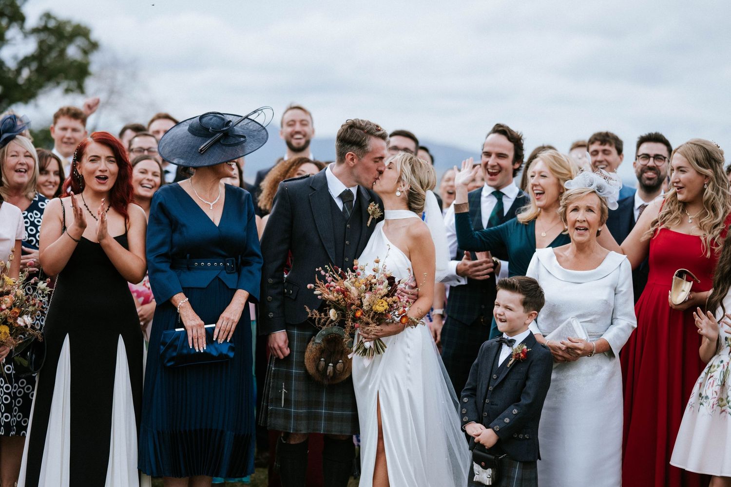 Wedding party group photo with guests in formal attire outdoors.