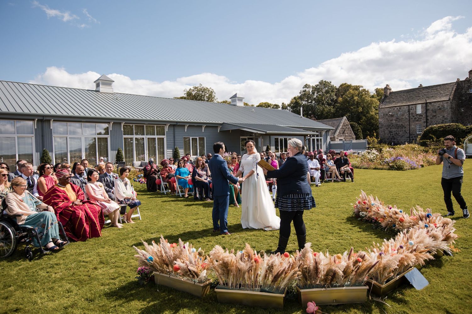 Outdoor wedding ceremony with guests seated on lawn and floral decorations in foreground.