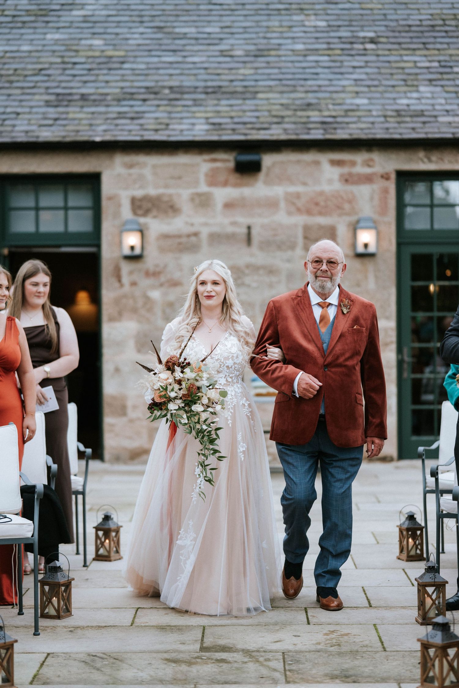 A wedding procession along a stone path with lanterns where a bride in blush gown walks with someone in a red blazer.