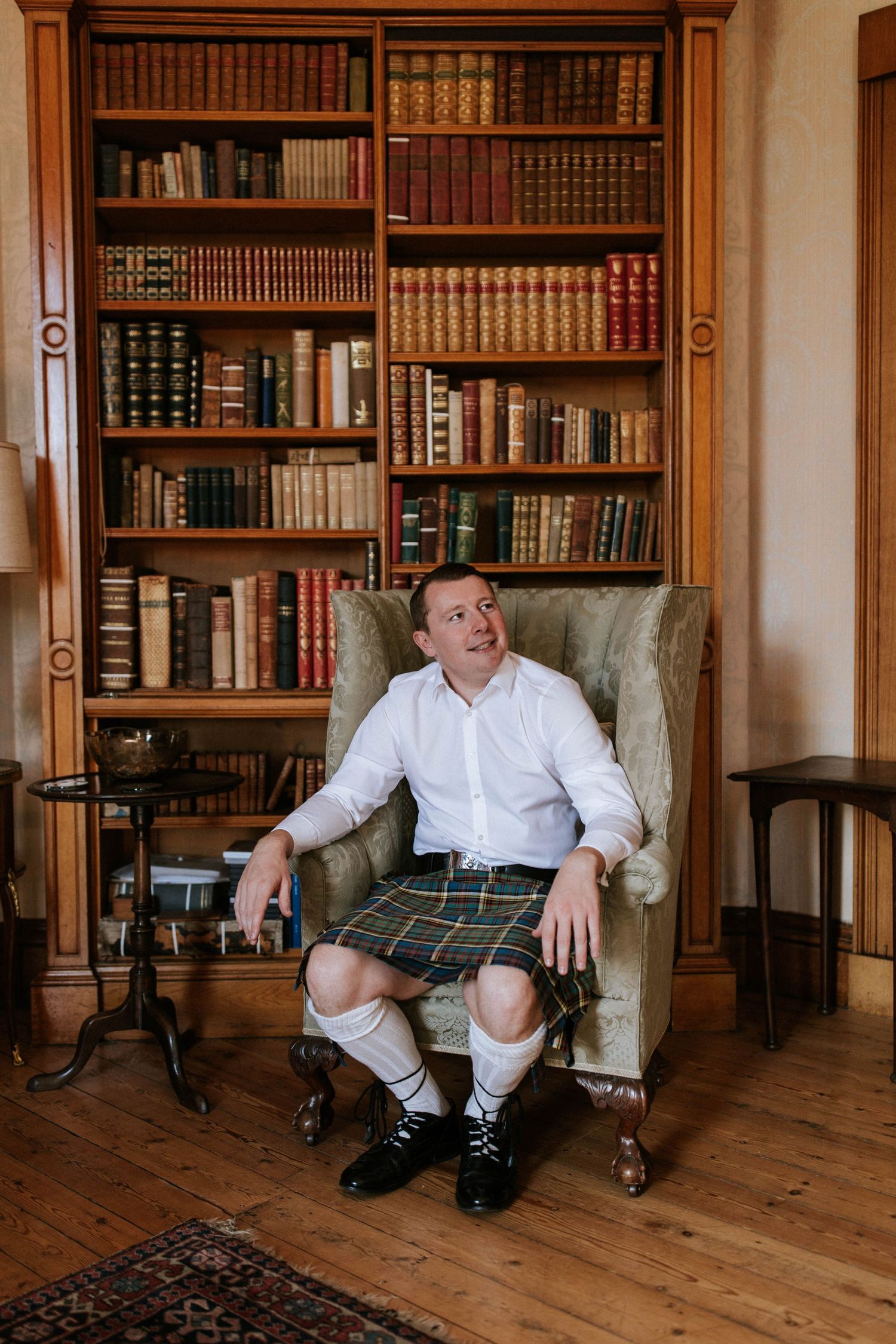 Person in traditional Scottish attire seated in library with wooden bookshelves.