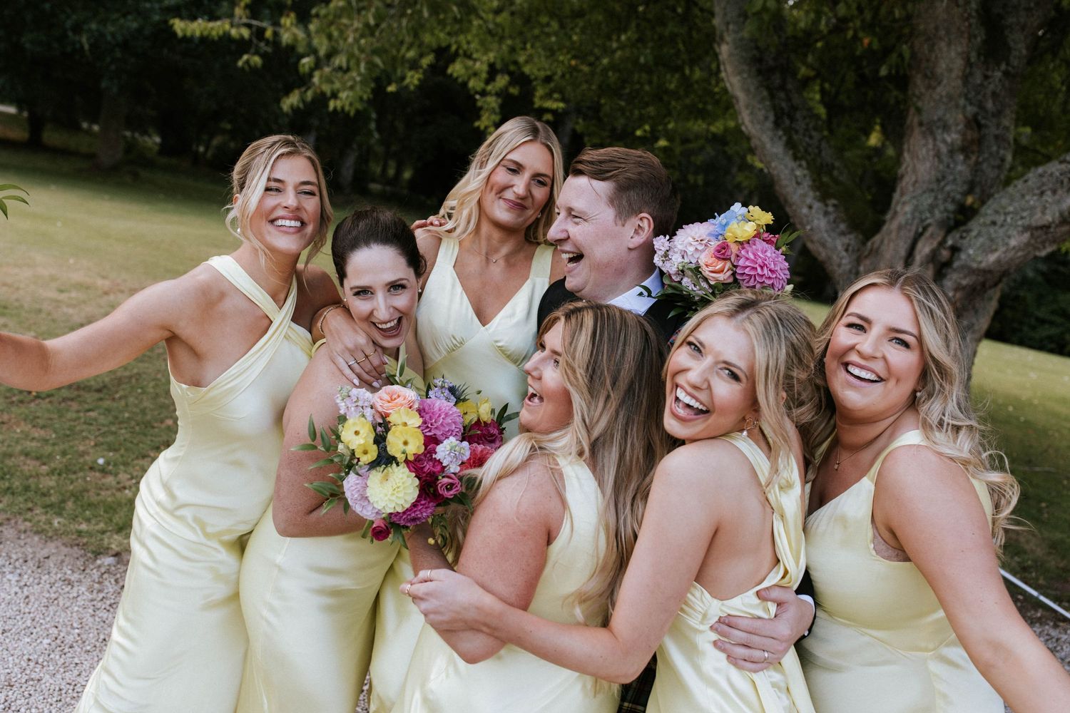 Bridesmaids in pale yellow dresses share a candid laughing moment on a garden path.