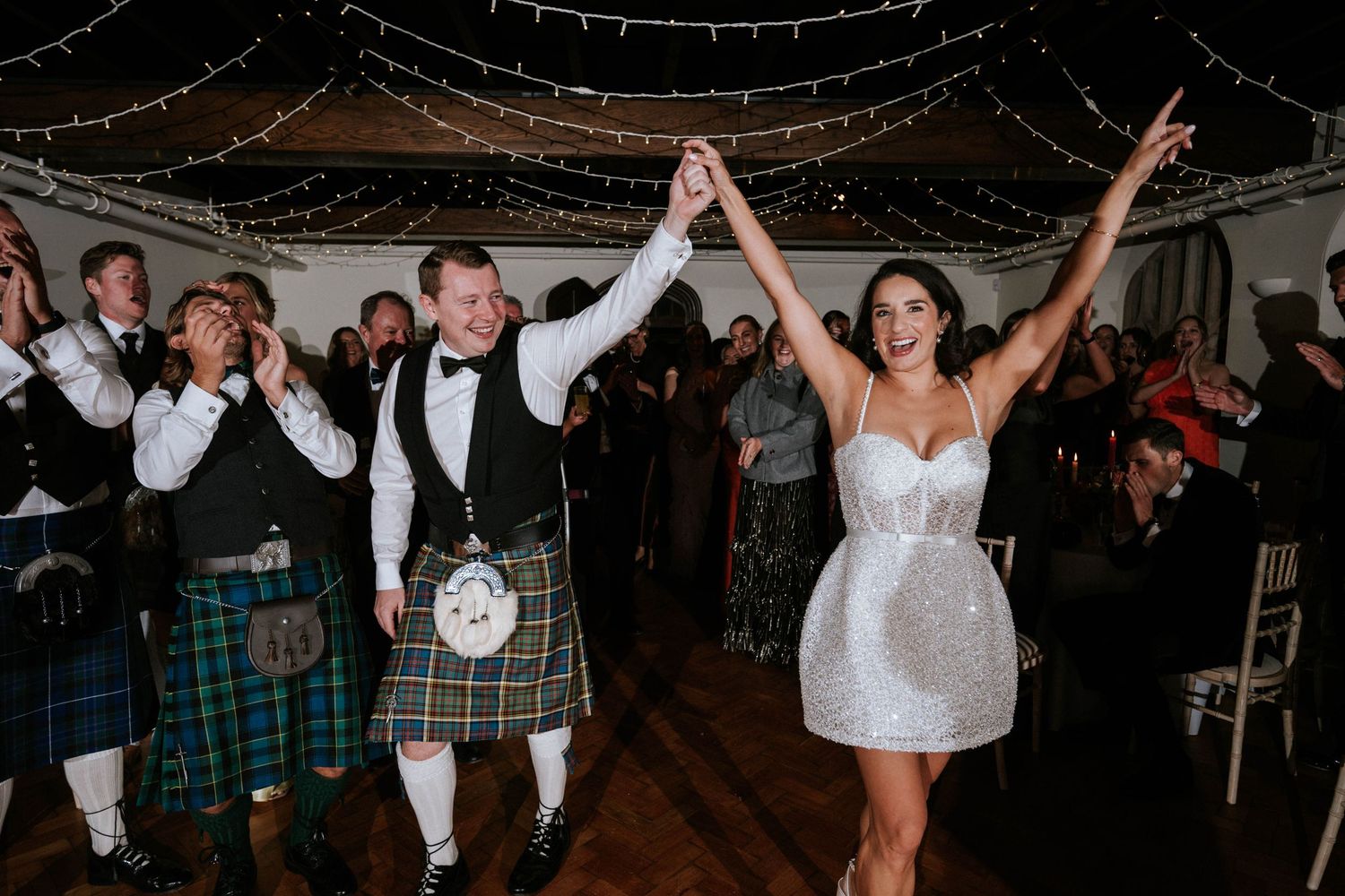 Wedding celebration with guests in kilts and couple dancing under string lights.