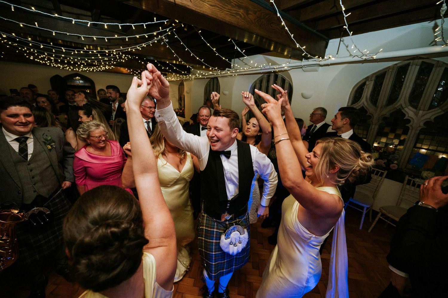 Energetic wedding party dancing and celebrating under decorative string lights.