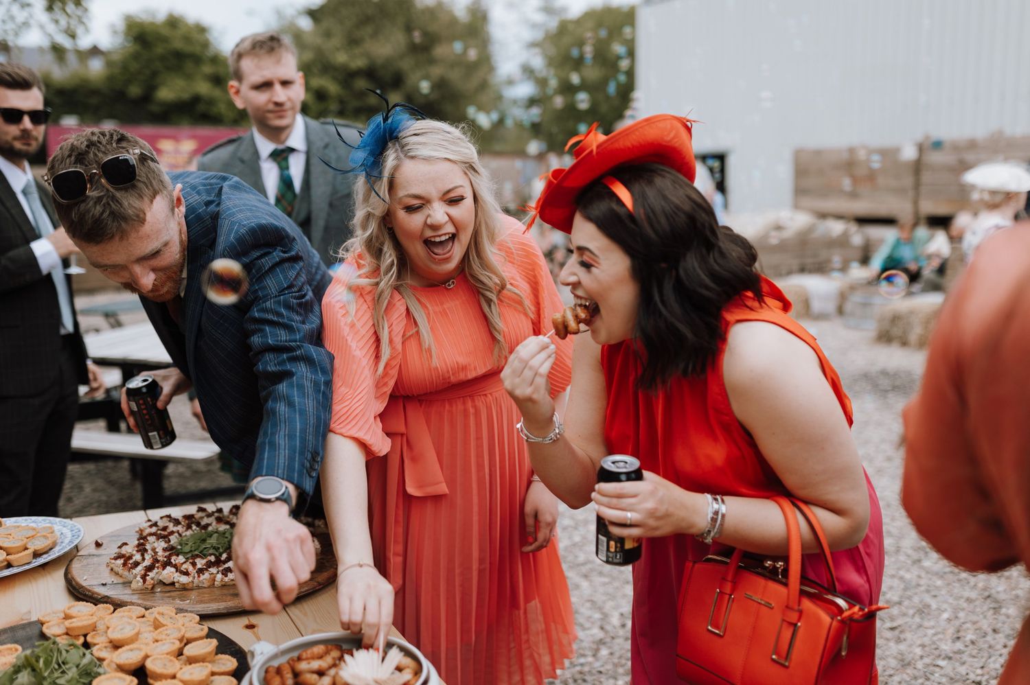 Wedding guests share joyful reactions while gathered around a food table in an outdoor celebration.