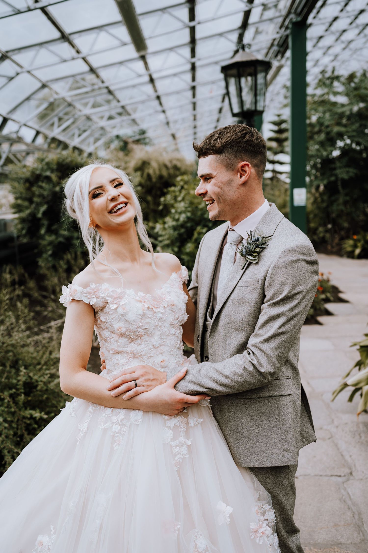 A joyful wedding couple laughs together in a sunlit greenhouse.