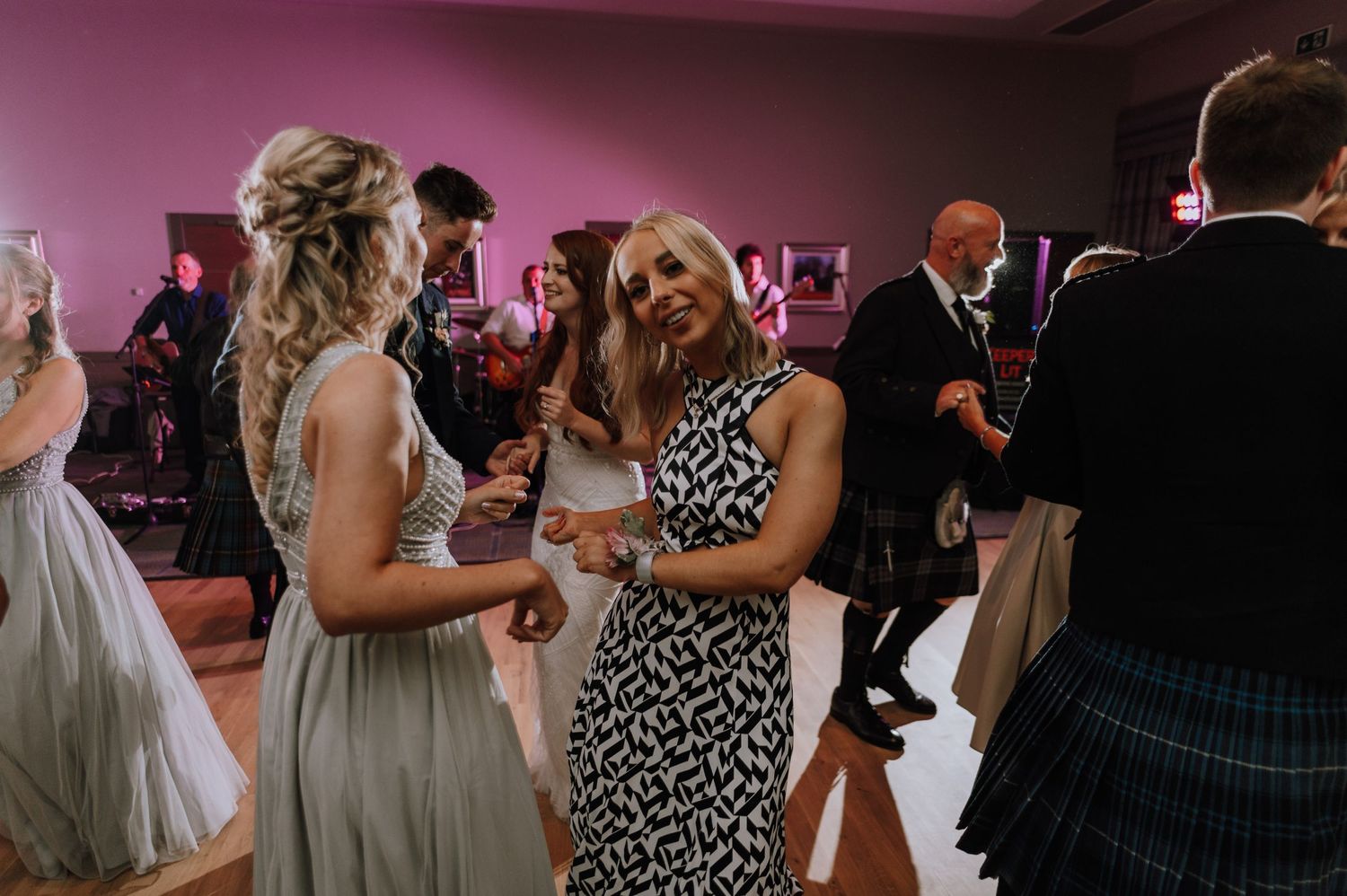 Guests in formal attire dance and celebrate on a reception dance floor with pink lighting.