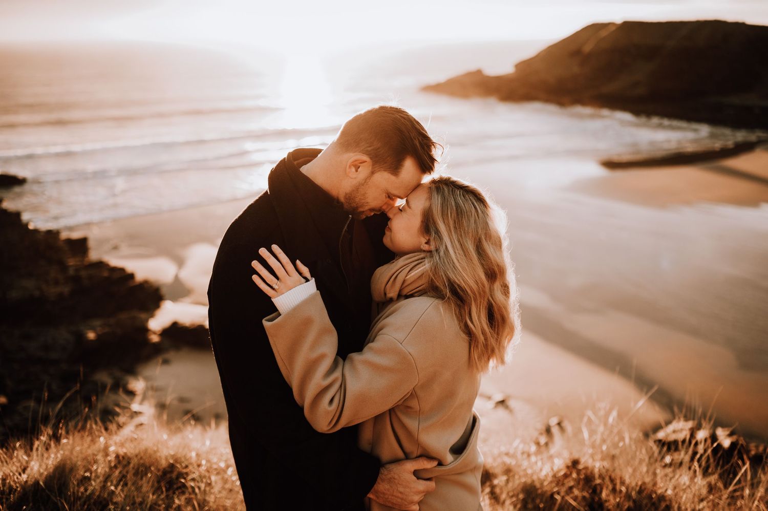 Couple shares tender moment against backdrop of dramatic coastal scenery.