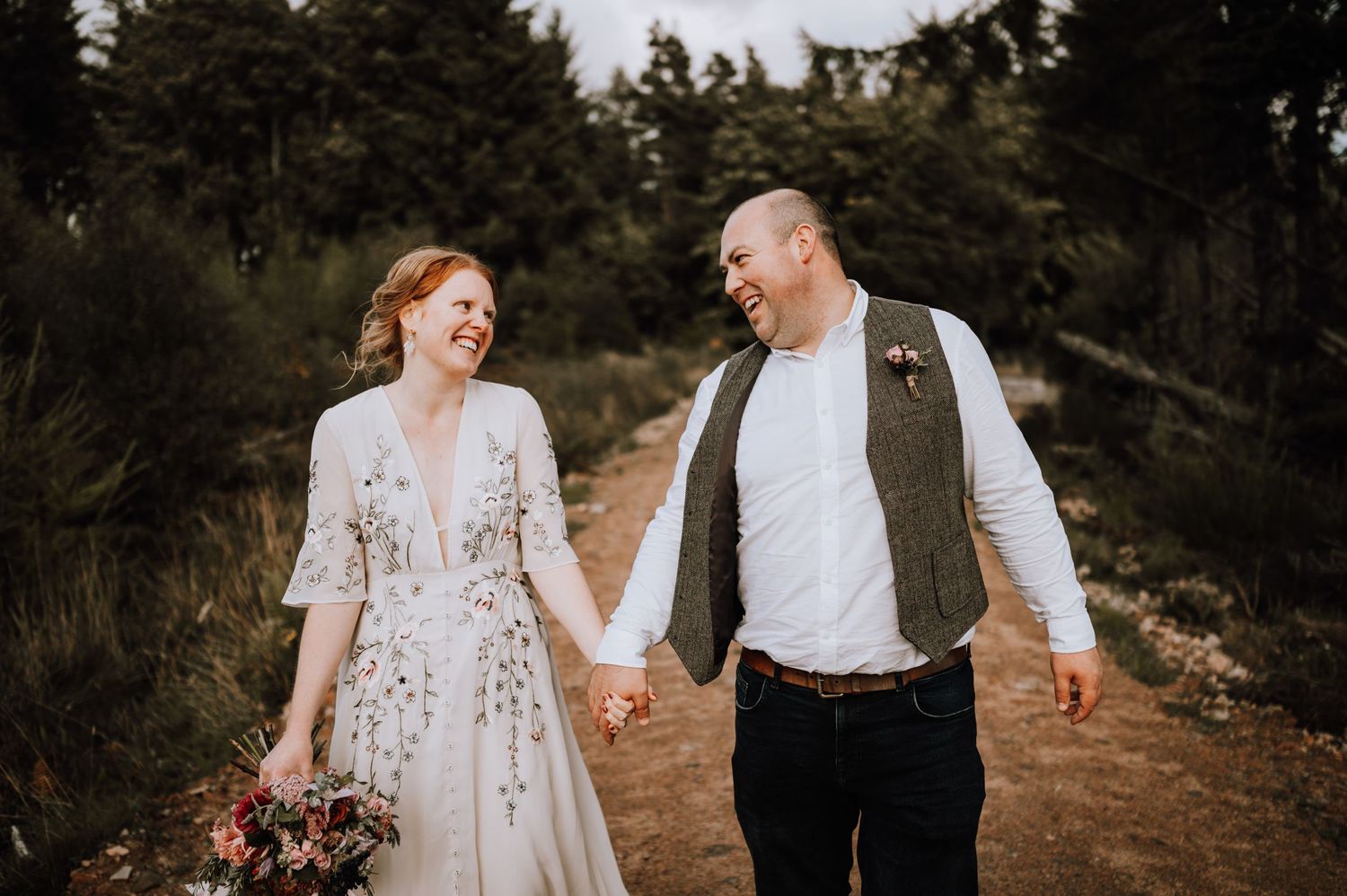 A joyful couple holding hands while walking on a dirt path surrounded by evergreen trees.