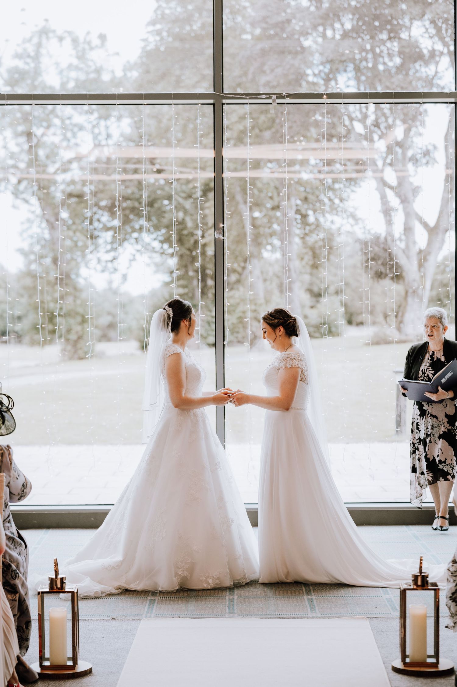 Two brides hold hands in matching white gowns against a bright window backdrop with lantern decorations.