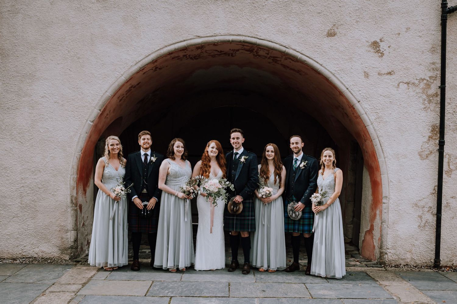 A wedding party poses together under an arched stone tunnel in formal attire.
