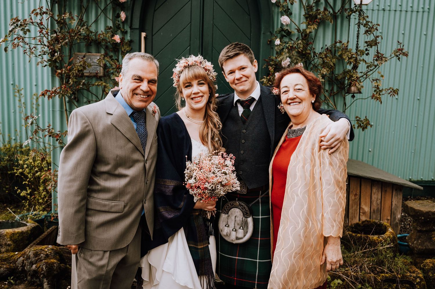 Small wedding group featuring traditional Scottish attire poses by rustic church entrance.
