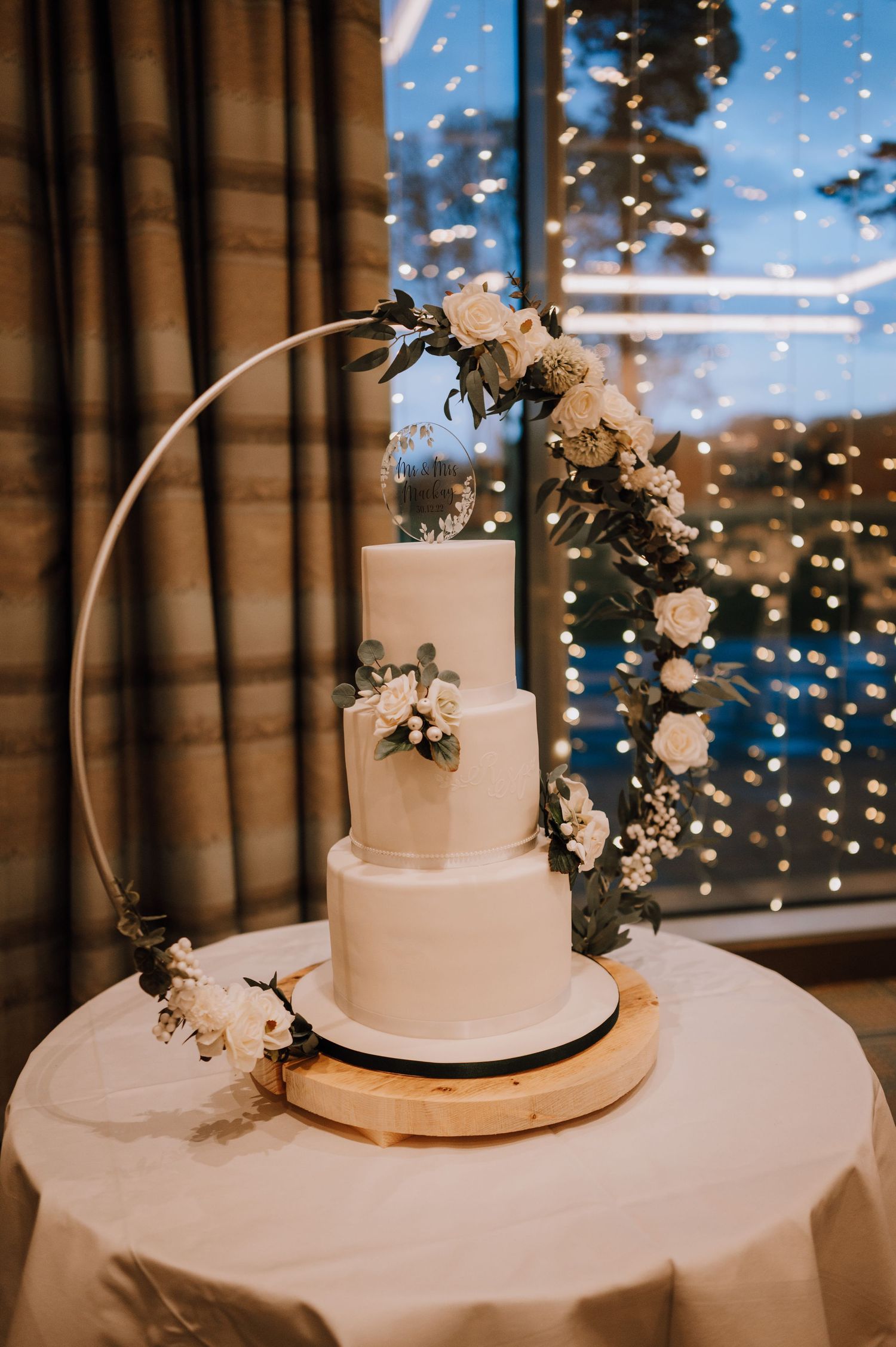 Elegant white wedding cake decorated with floral arch against twinkling light background.