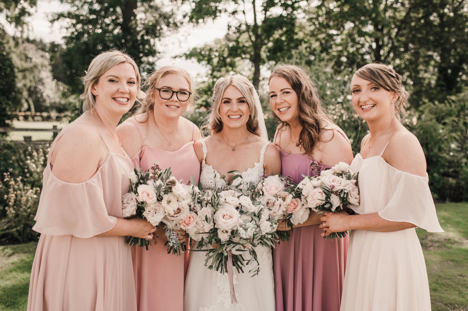 Bridal party in soft pink and mauve dresses holds floral bouquets while posing outdoors.