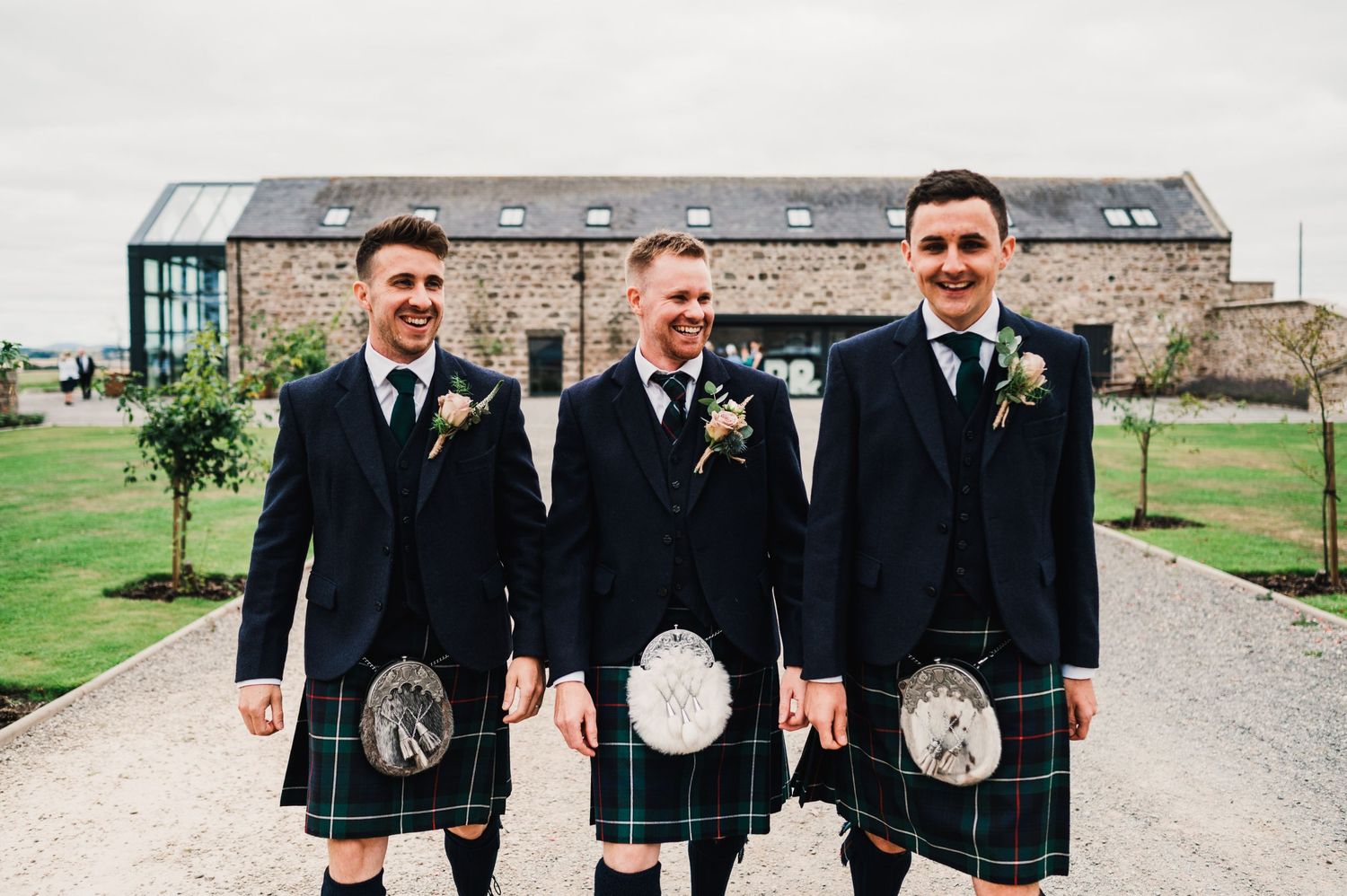 Three men in traditional Scottish wedding attire pose on a path near a stone building.