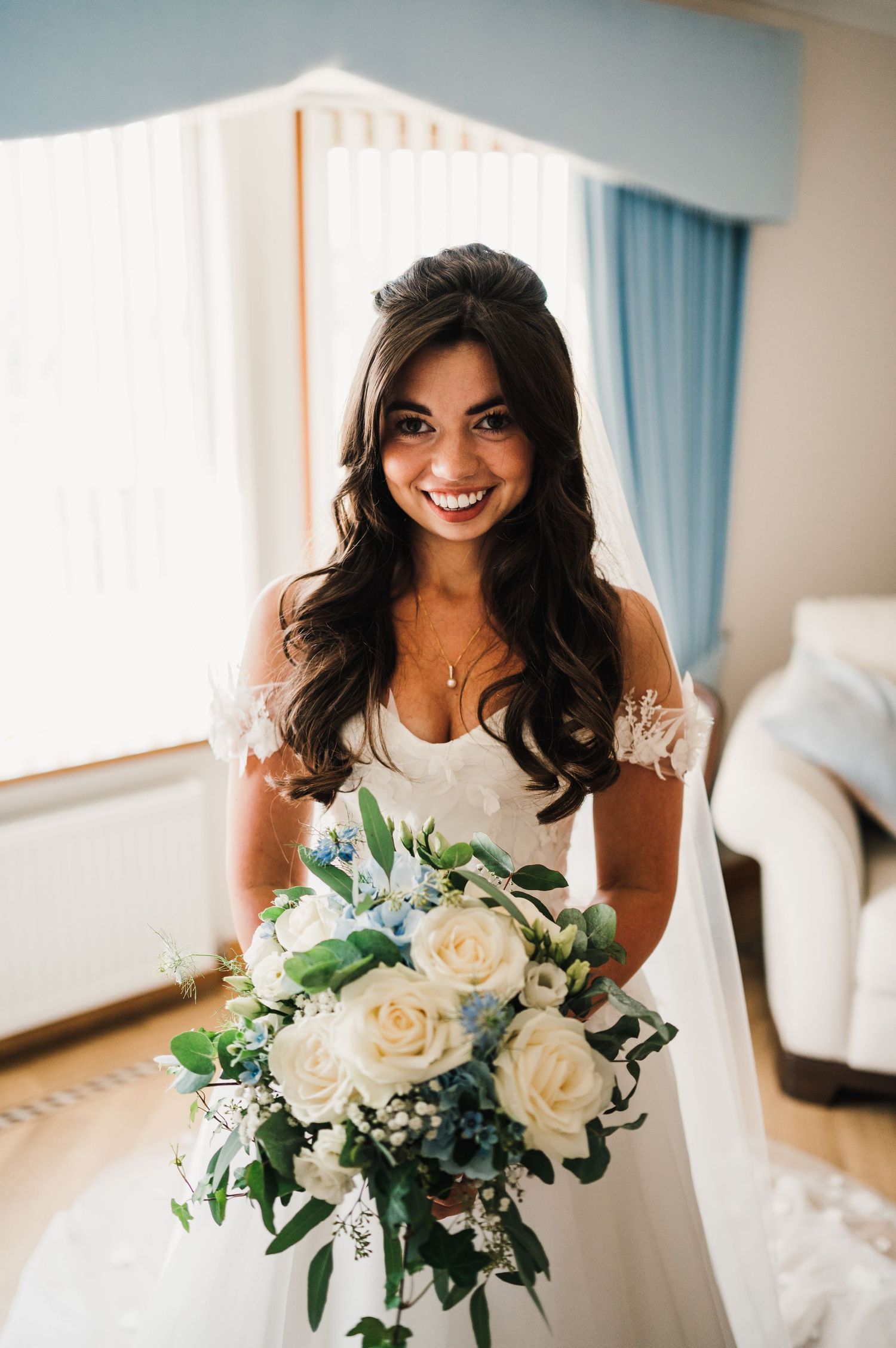 A bride holds a white rose bouquet while standing in a bright room with blue curtains.