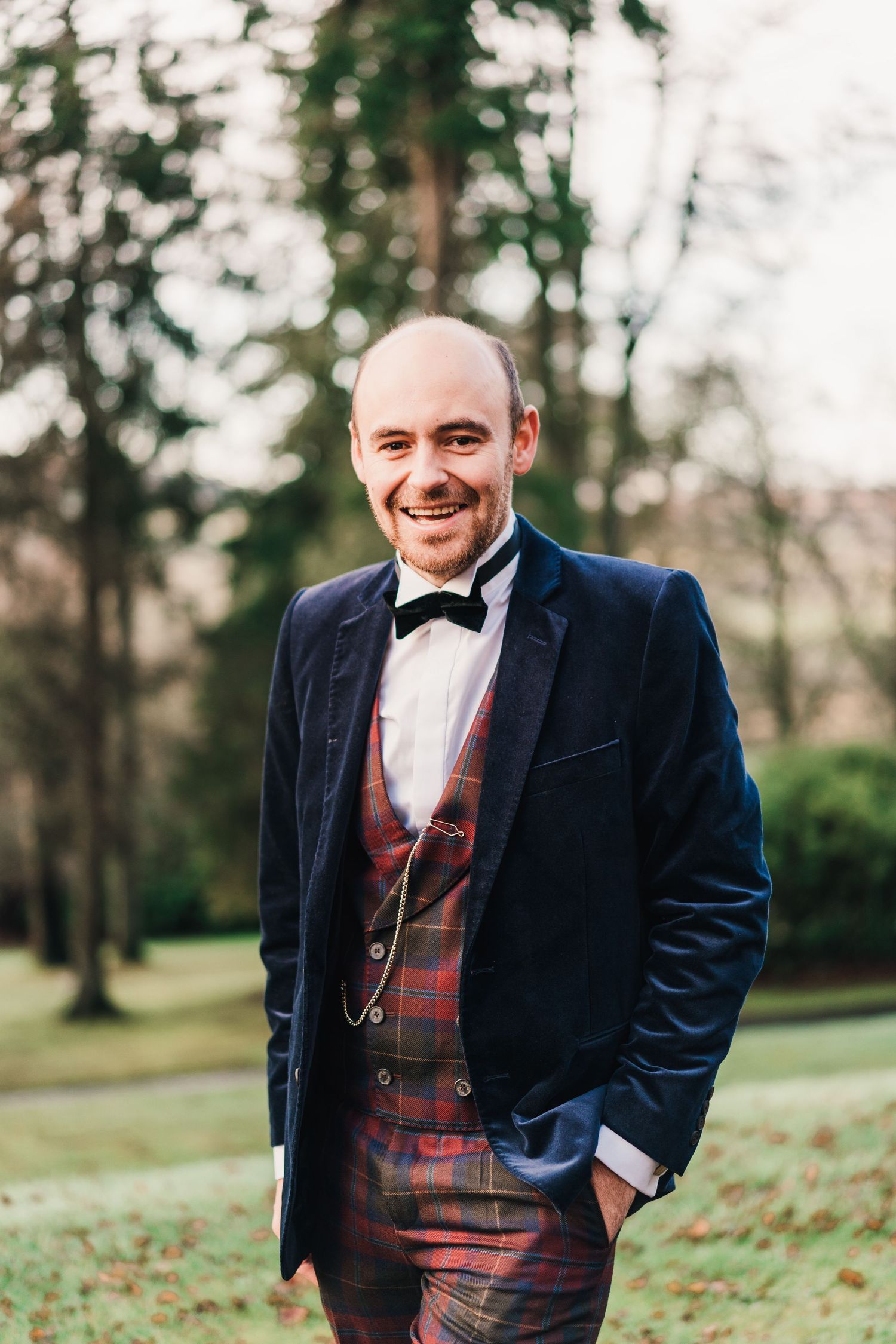 Groom in navy velvet jacket and plaid vest poses outdoors in a park setting.