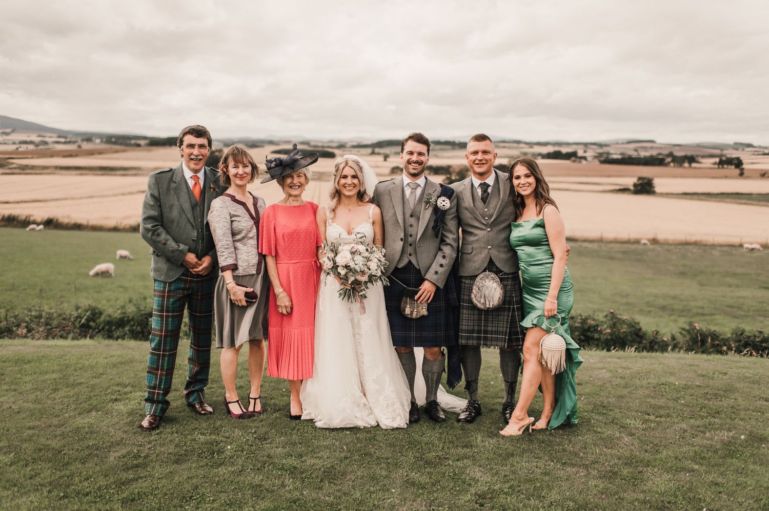 Wedding party in mix of formal wear and traditional Scottish kilts pose on hillside.