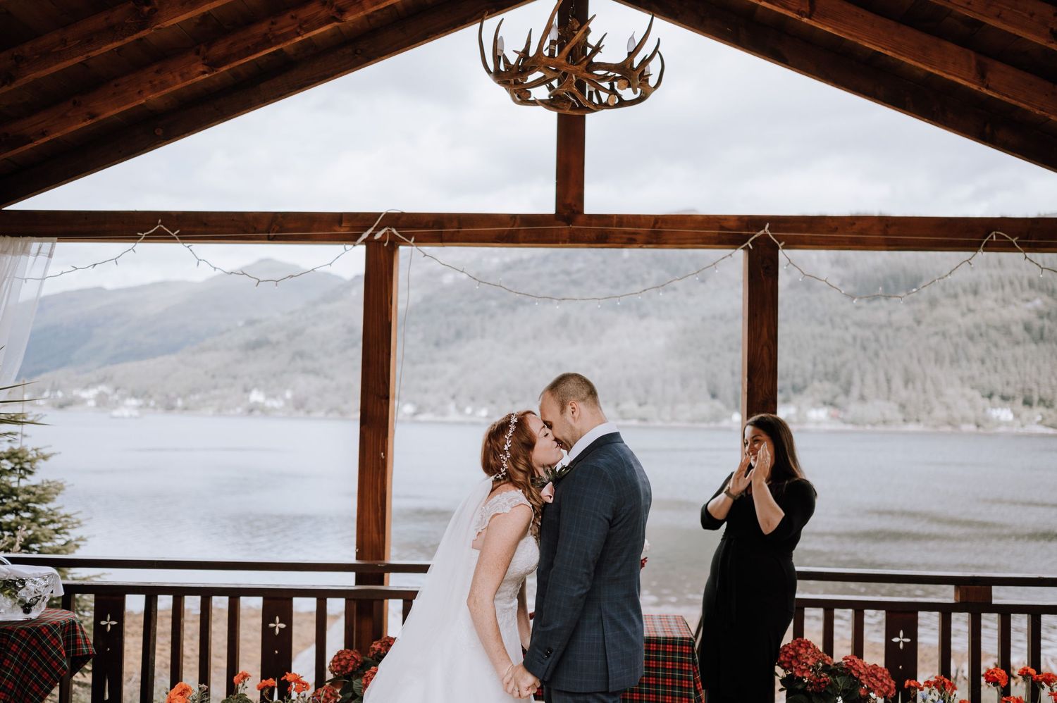 Wedding ceremony moment under rustic pavilion with antler chandelier and mountain lake backdrop.